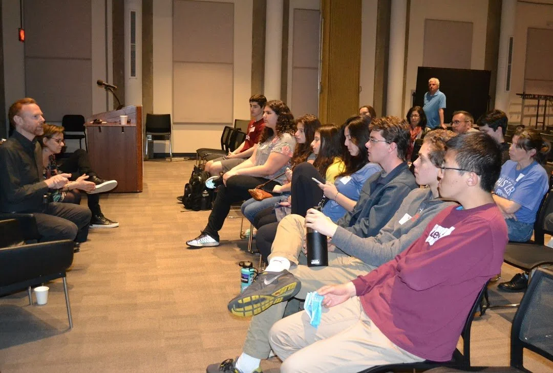 A group of young adults attending a presentation or lecture in a conference room, seated in rows facing a speaker.