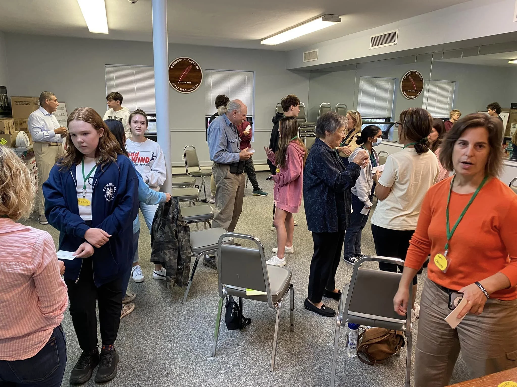 A group of people, including children and adults, are gathered in a room that appears to be a community center or meeting room. Some people are standing in line, talking, or looking at papers. The room has chairs, boxes, and a clock on the wall.