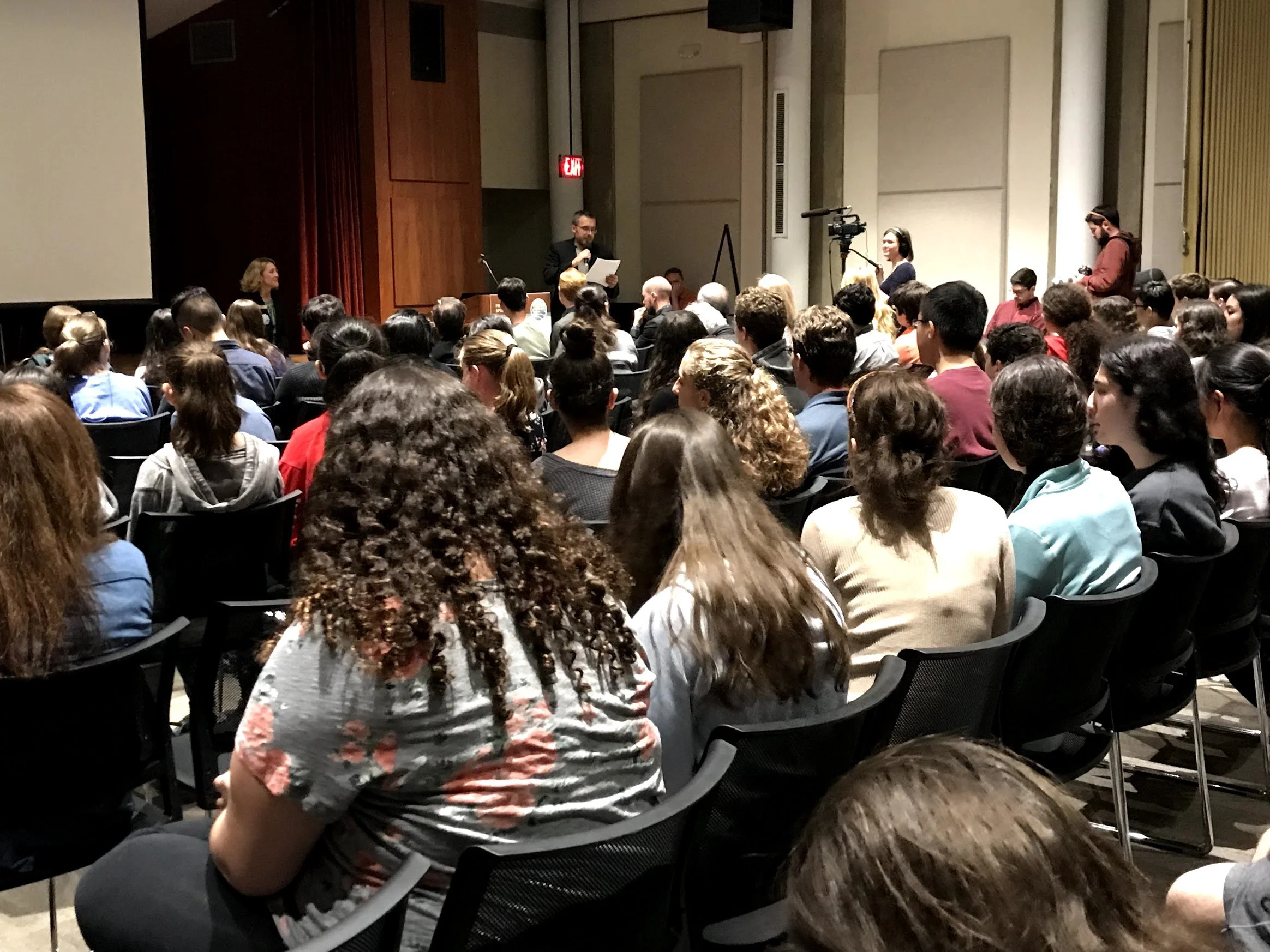An audience of diverse people seated in chairs facing a stage in a conference room, with a man standing on stage reading from a paper, while a woman on the left looks on and a camerawoman films the event.
