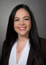 A woman with long black hair, wearing a white blouse and smiling, posed against a dark background.