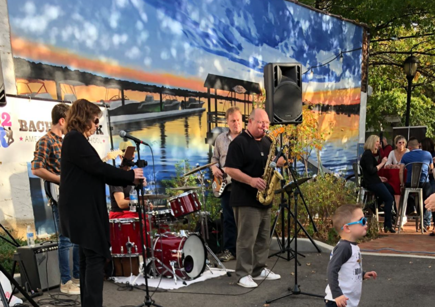 A live band performing outdoors at sunset with a cityscape in the background, featuring a drummer, saxophonist, guitarist, and singer, with audience members seated at tables and a young child in sunglasses watching the performance.