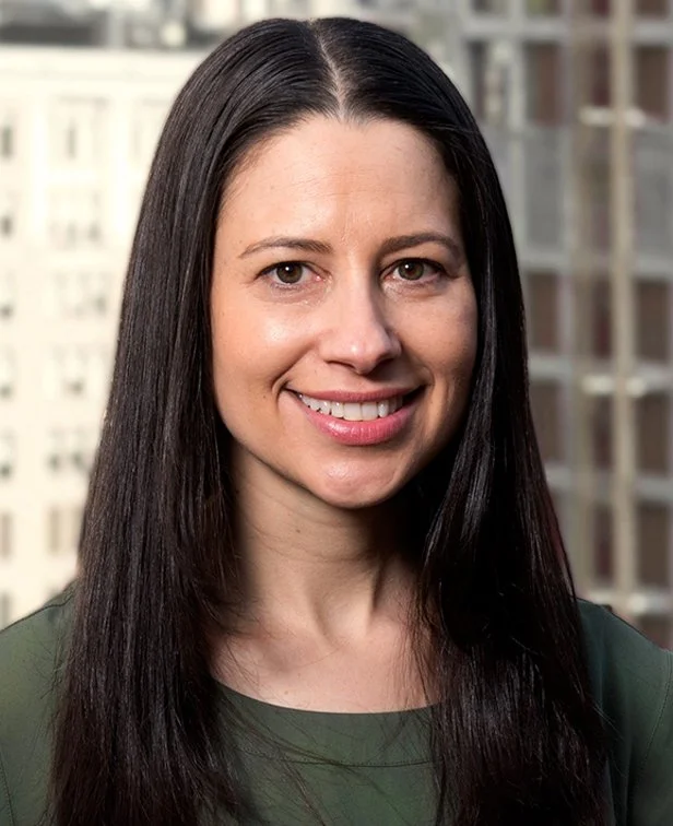 A woman with long straight dark hair smiling outdoors with a cityscape of tall buildings in the background.