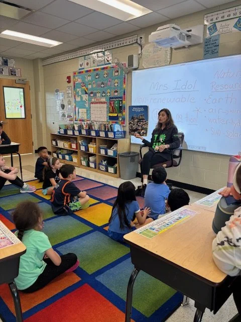 Elementary classroom with students sitting on colorful carpet squares, listening to a teacher who is reading a book. Whiteboard displays notes about renewable resources like earth, water, and sunlight.
