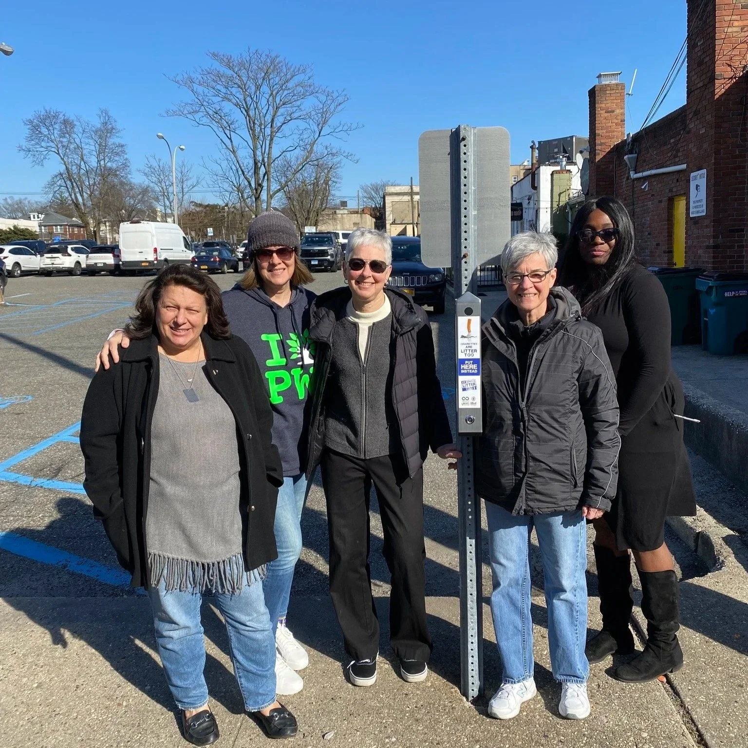 Five women standing together outdoors near a parking lot, smiling, some with their arms around each other. They are in front of a parking meter or payment station on a city street with cars, trees, and brick buildings in the background.