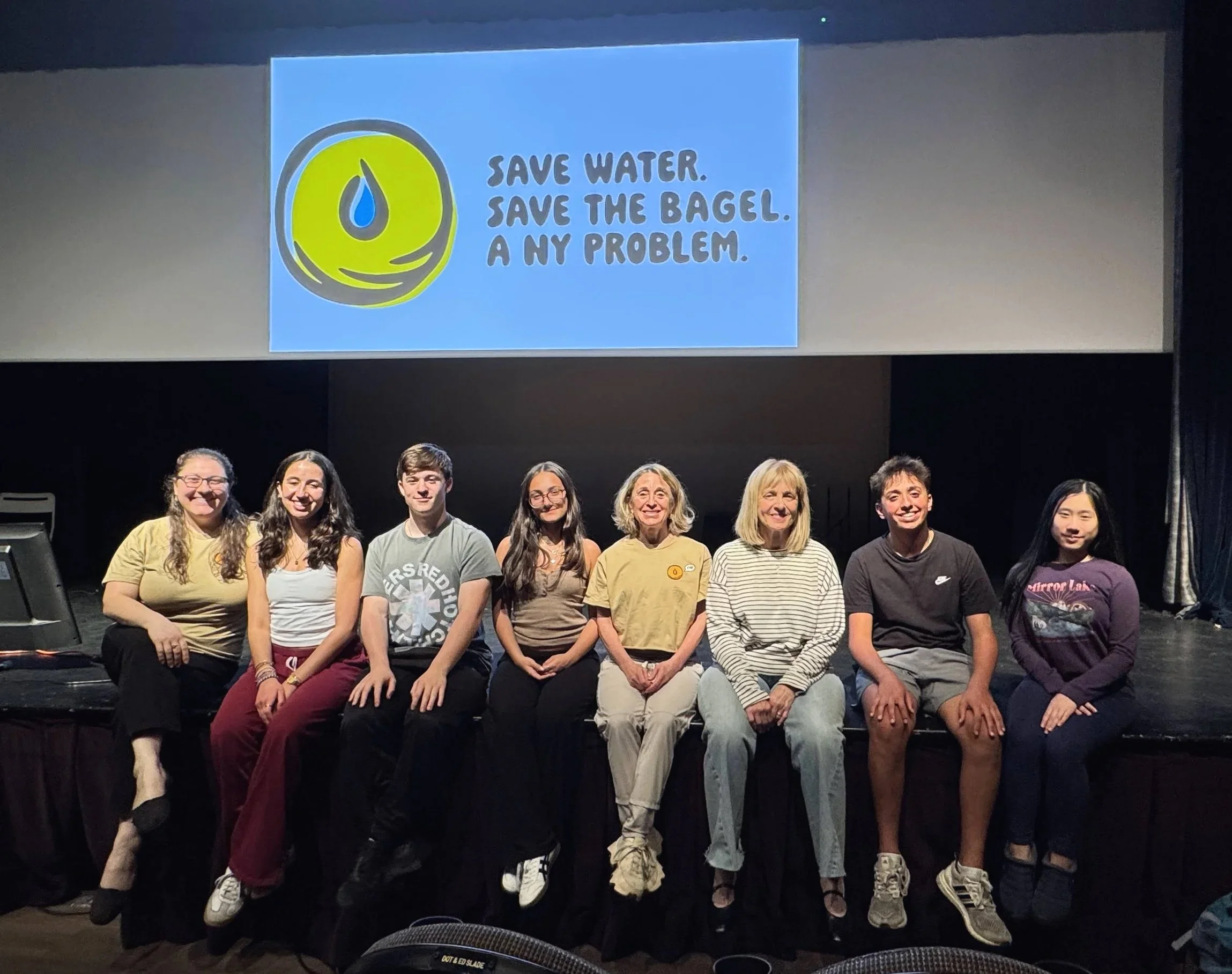 Group of eight people sitting on a stage in front of a large screen displaying a cartoon bagel with a water droplet and the text 'Save water. Save the bagel. A NY problem.'.
