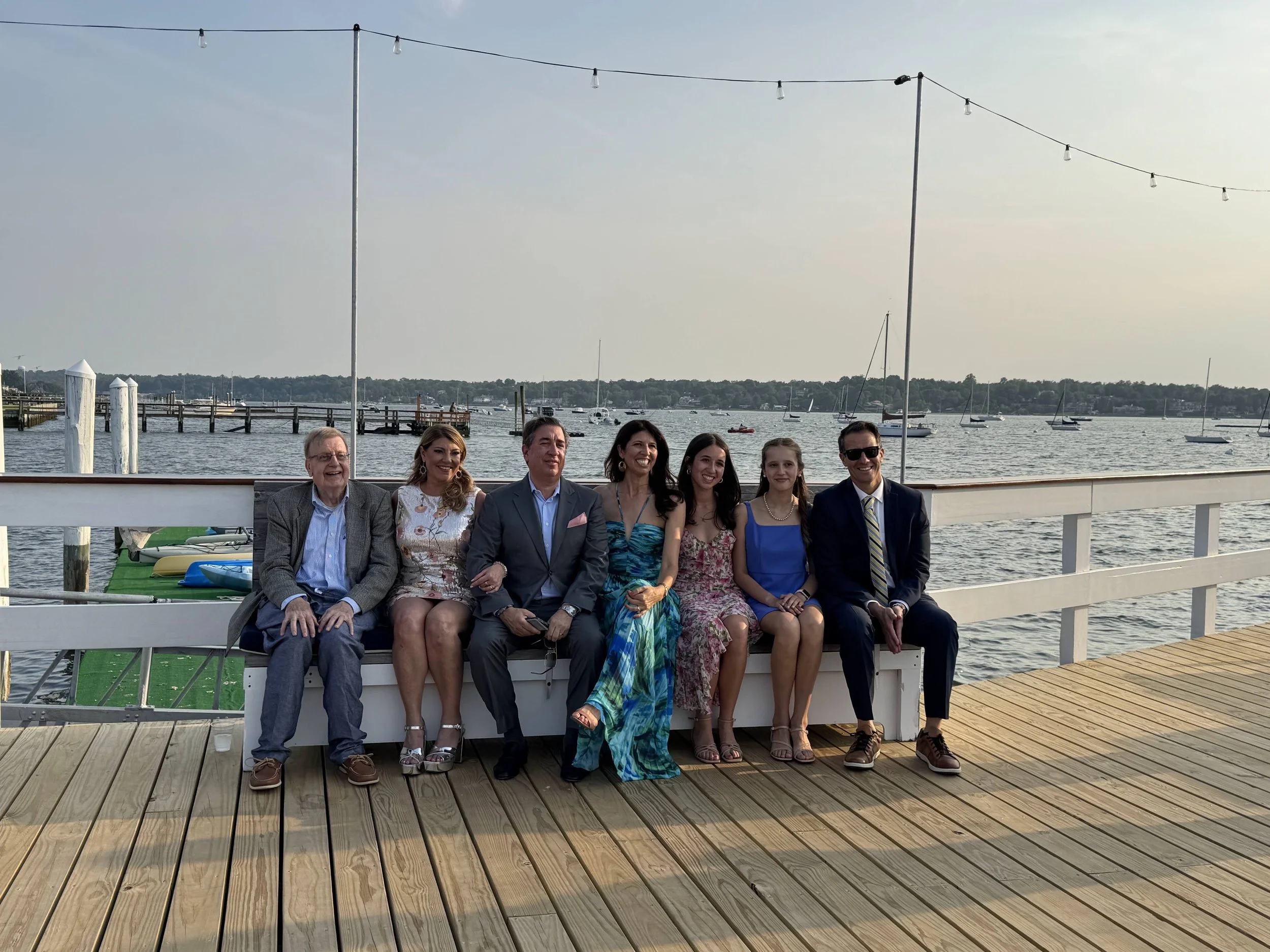 Seven people sitting on a bench on a dock by a body of water, smiling and wearing semi-formal clothing, with sailboats in the background and string lights overhead.