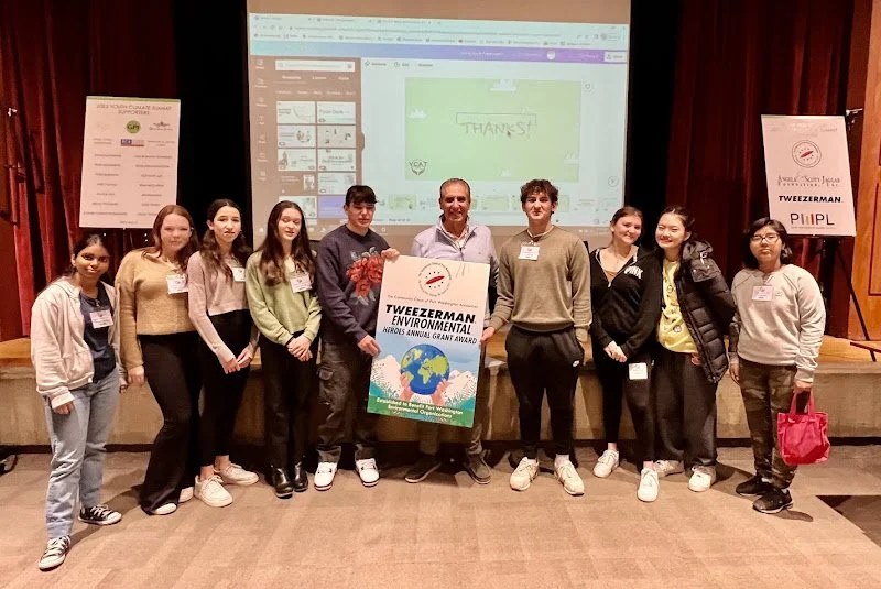 A group of nine young people and one adult standing on a stage, holding a sign that reads "TWEZZERMAN ENVIRONMENTAL MARS ANNUAL GRANT AWARD." They are smiling and posing for a photo, with a large screen in the background displaying a presentation sli