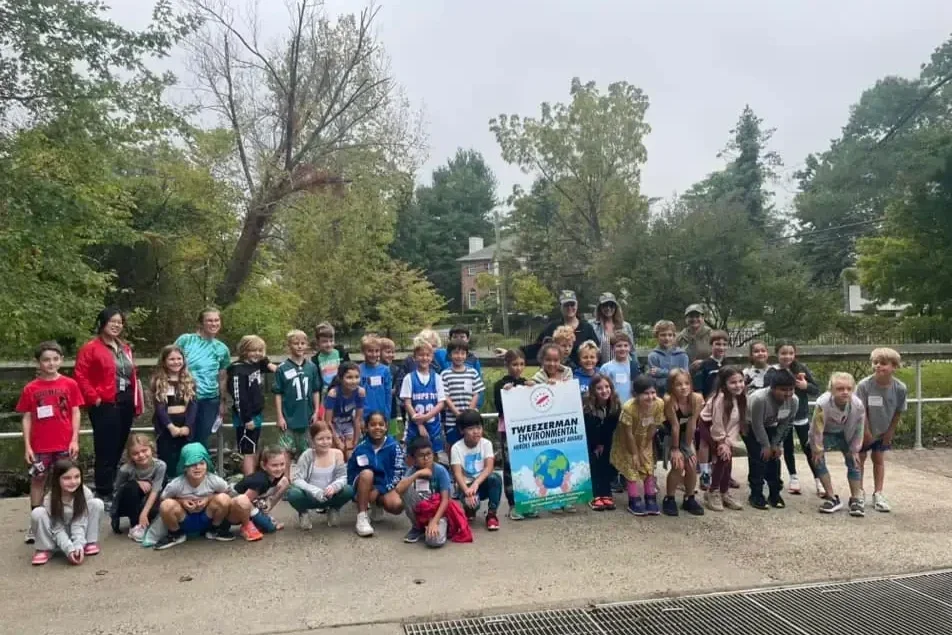 Group of children and adults outdoors holding a sign that reads 'Tweezerman Environmental Ambassador Global Climate Change'.
