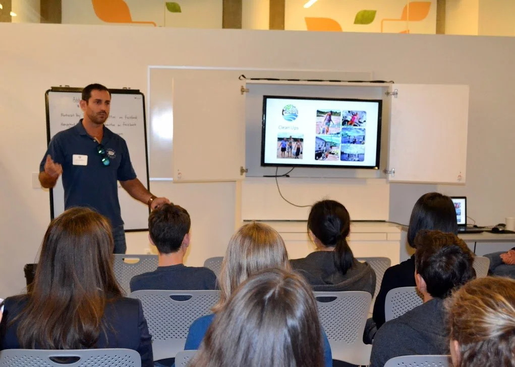 A man giving a presentation to a group of people in a classroom setting. The man is standing near a whiteboard and a screen displaying images. The audience is seated in chairs facing him.