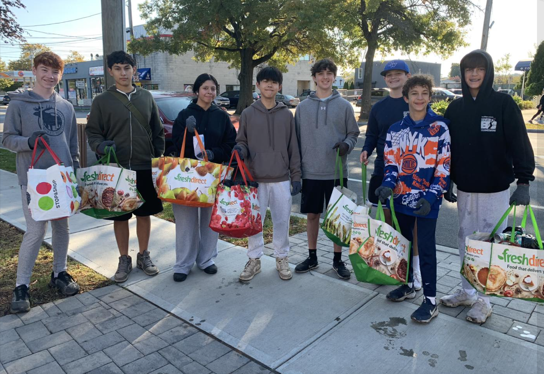 A group of nine young teenagers standing on a sidewalk outdoors, holding shopping bags from FreshDirect, wearing casual and sporty clothing, with trees and a parking lot in the background.