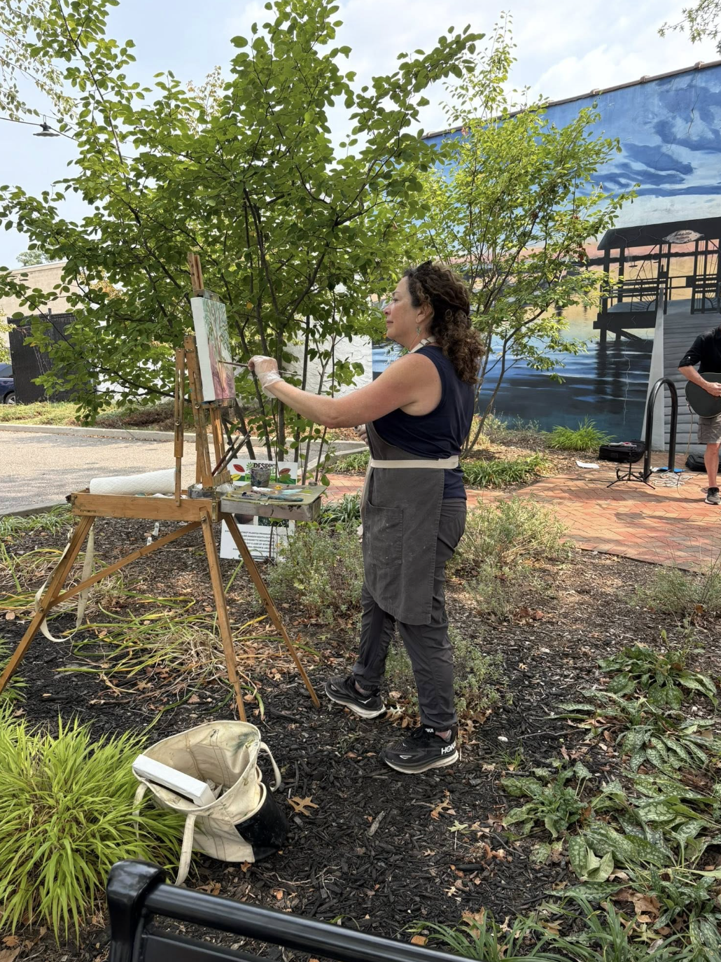A woman painting outdoors on an easel, surrounded by greenery, with a mural of a pier and water in the background.