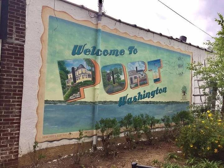 Colorful mural on an exterior wall welcoming visitors to Port, Washington, featuring images of local buildings and scenic waterfront, with small bushes and plants in front.