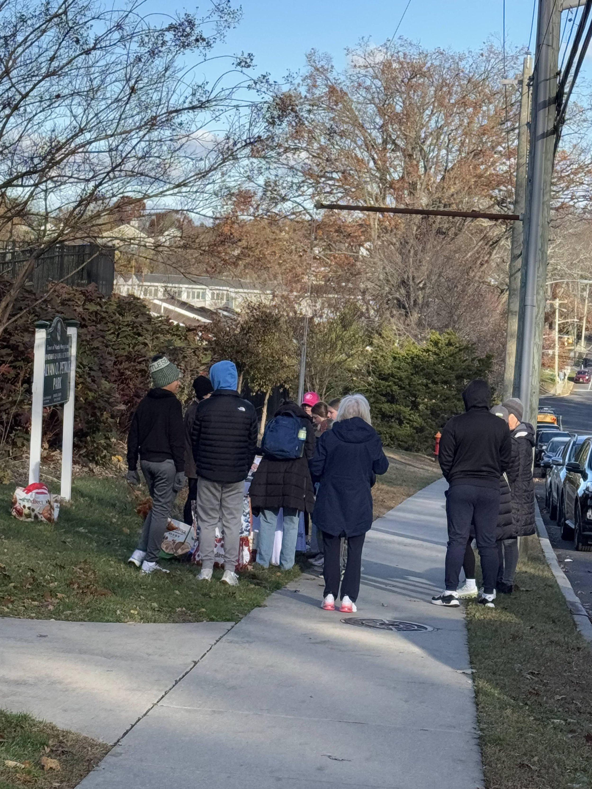 A group of people gathered on a sidewalk near trees with autumn foliage, some carrying bags and wearing warm clothing, possibly waiting in line.