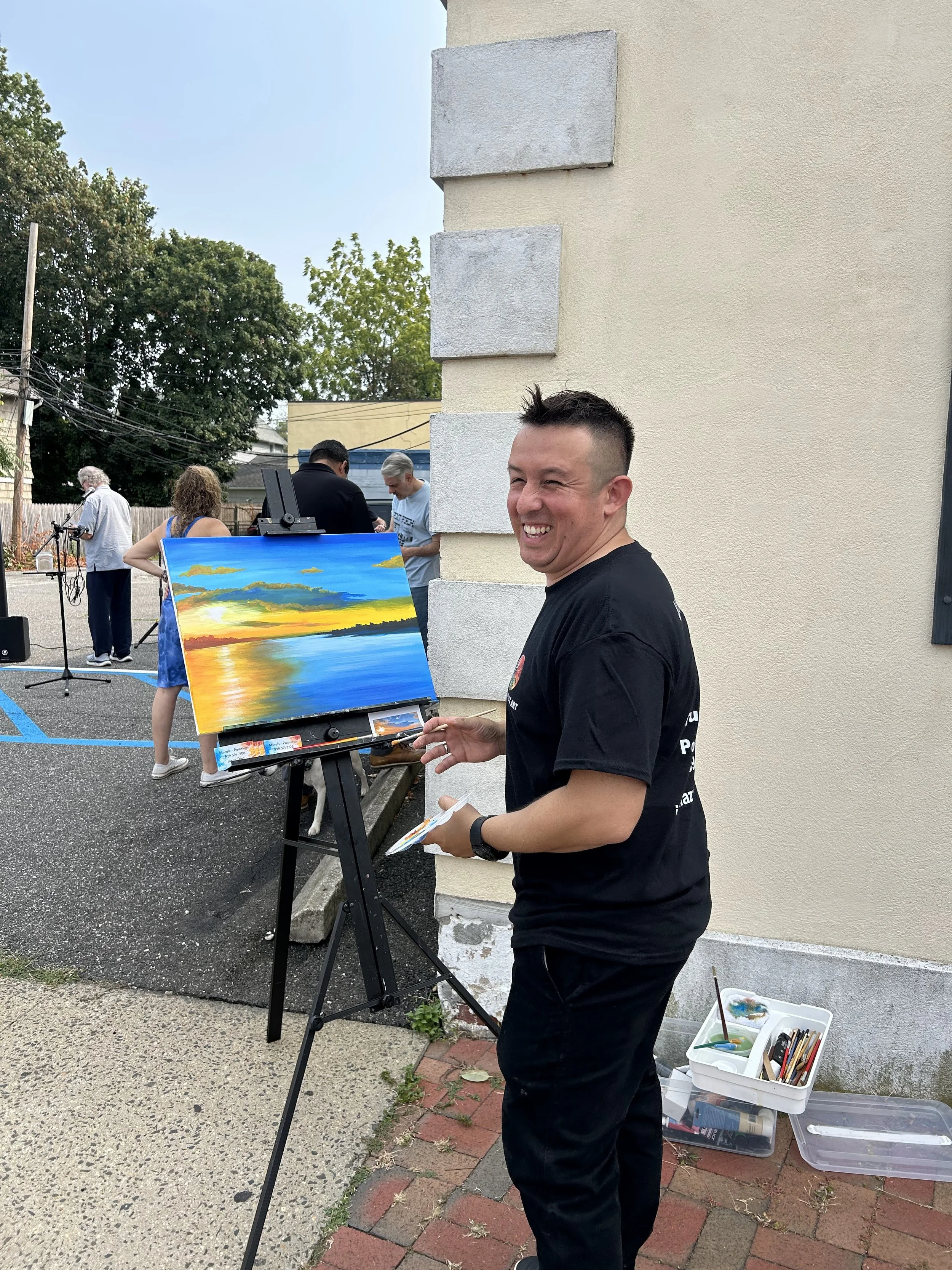 A man painting a sunset landscape on a small canvas outdoors, with people and a musician in the background.