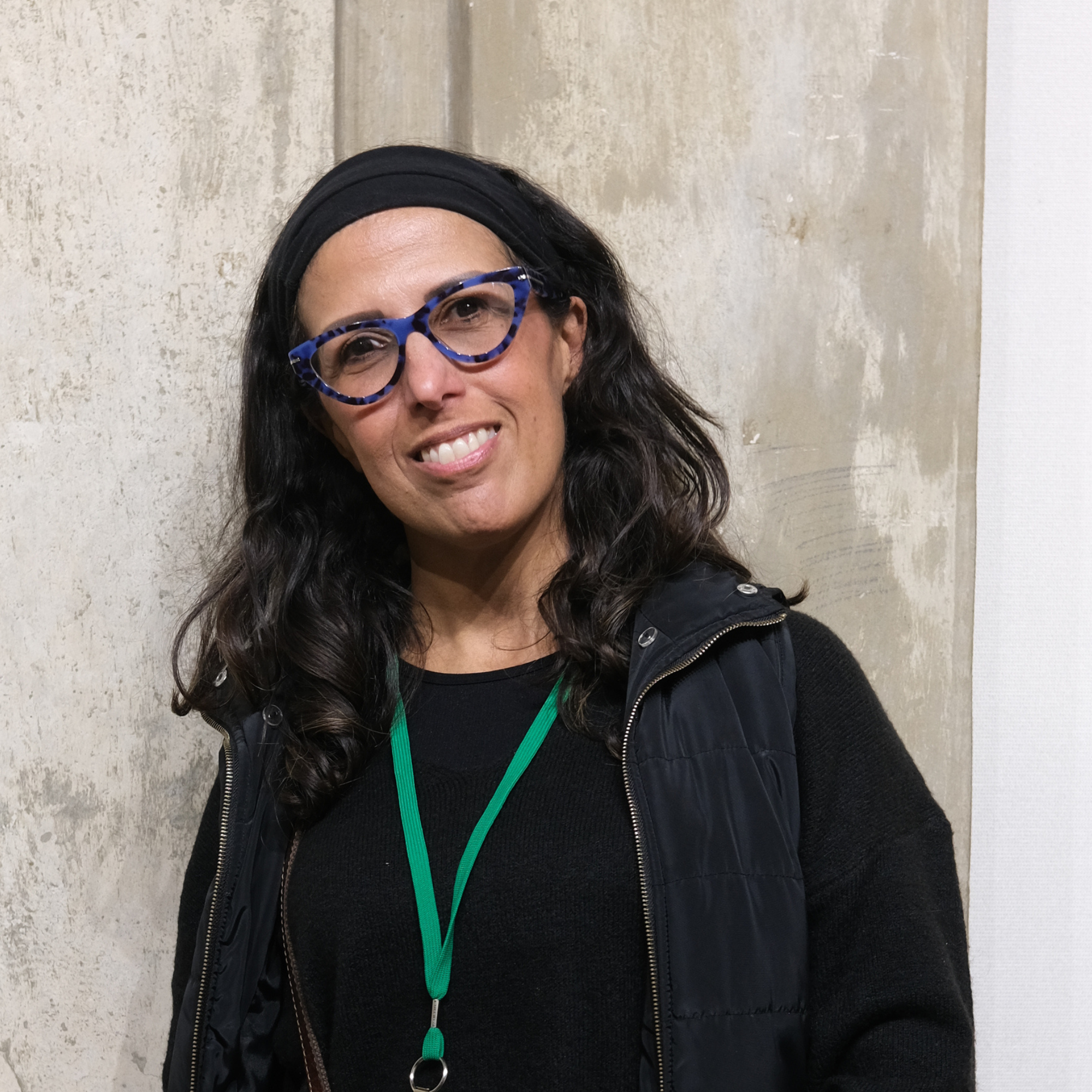 A woman with long dark wavy hair wearing blue patterned glasses, a black headband, a black shirt, and a black vest, smiling in front of a textured beige wall.