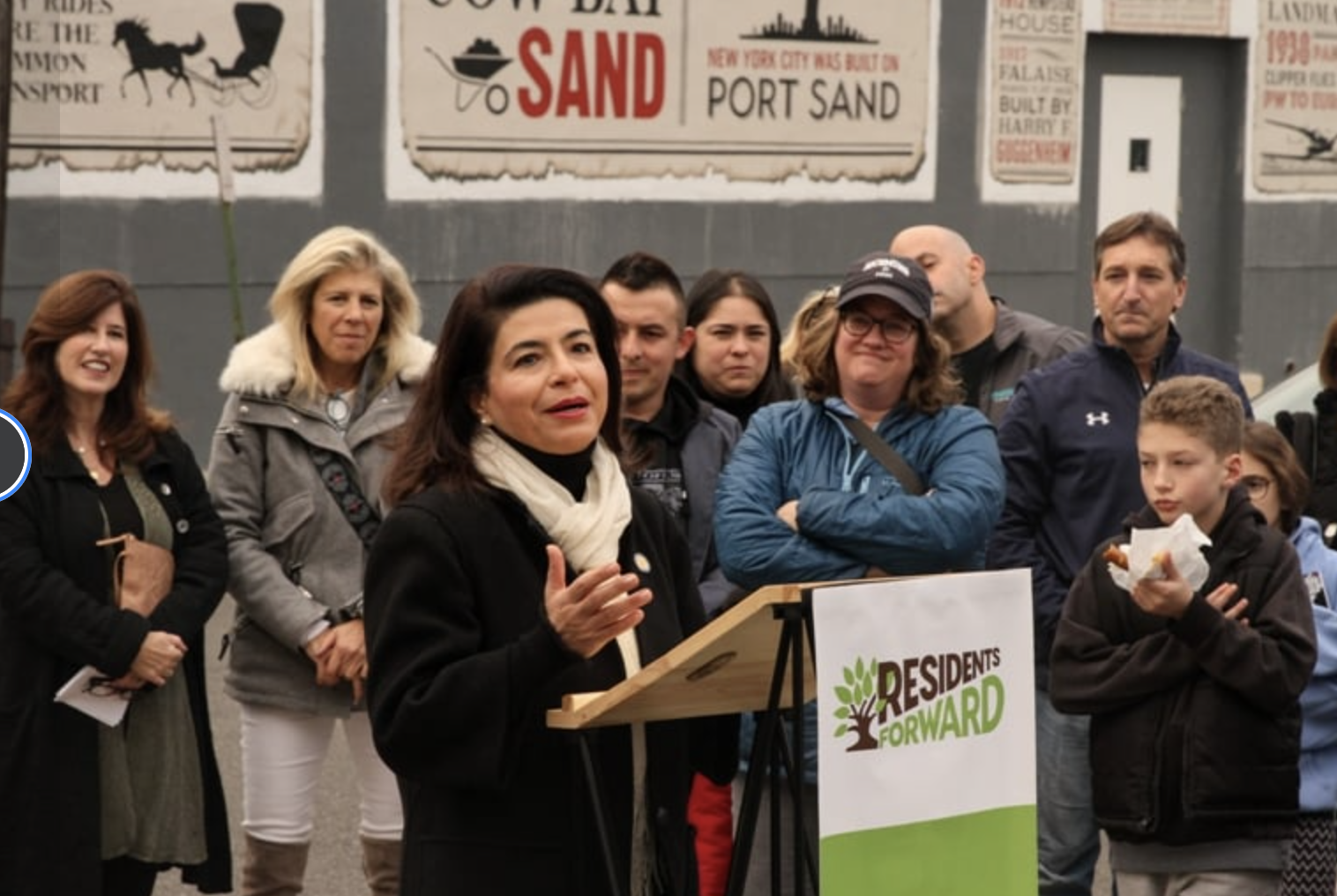A woman speaking at a podium outdoors, surrounded by a diverse group of people listening, with signs about sand and a political sign that says 'Residents Forward' in the foreground.