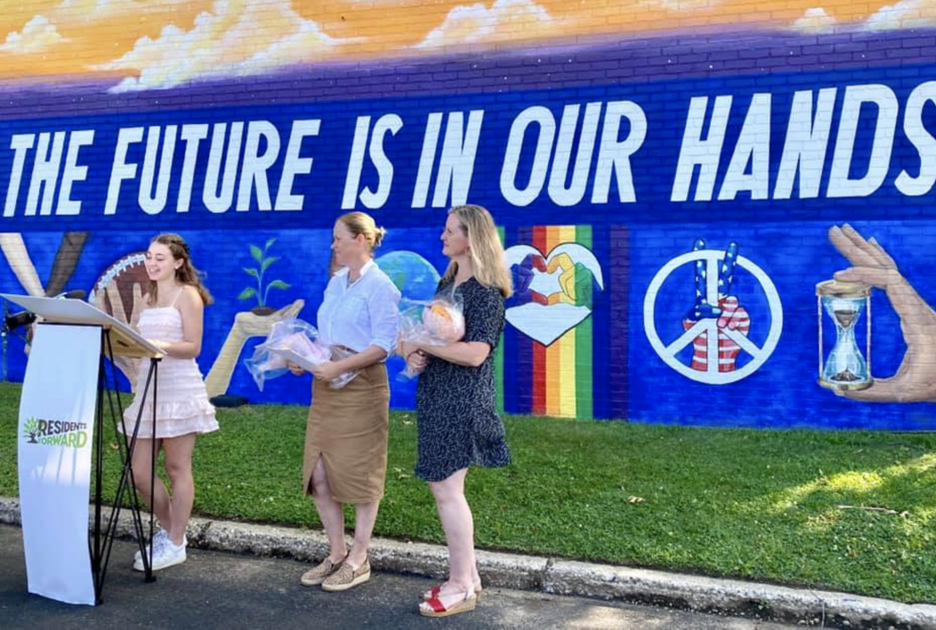 Three women and a girl standing in front of a wall with a mural and large text that says 'The future is in our hands.' The mural features symbols of a rainbow flag, a heart, a peace sign, a hand holding an hourglass, and environmental images. One wom