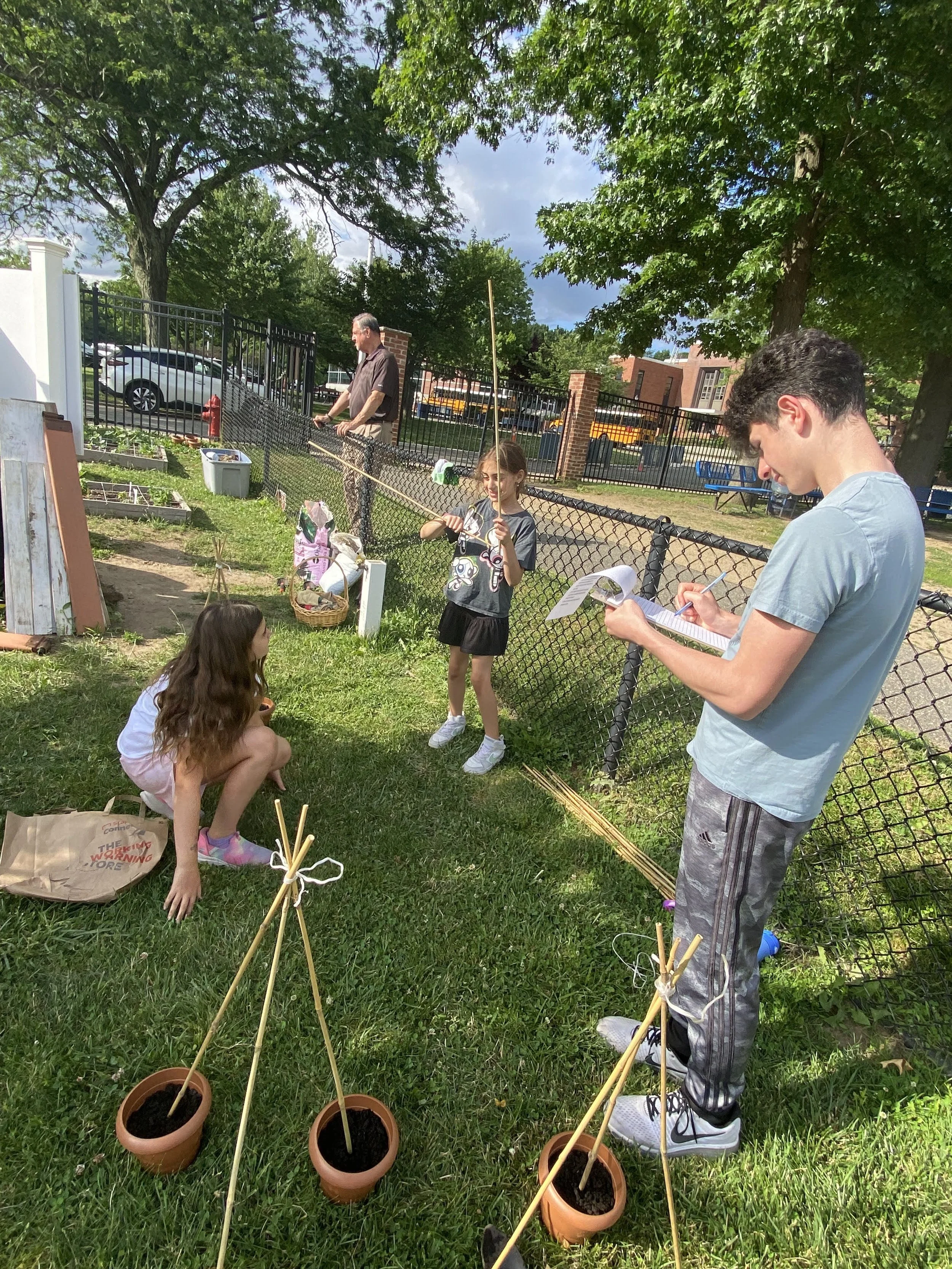 Two young girls and two young boys in a backyard with a fenced area, some gardening supplies, and potted plants. One girl is crouching down, and another girl is standing, holding a fishing pole. One boy is taking notes on a clipboard, and another man is working in the garden in the background.