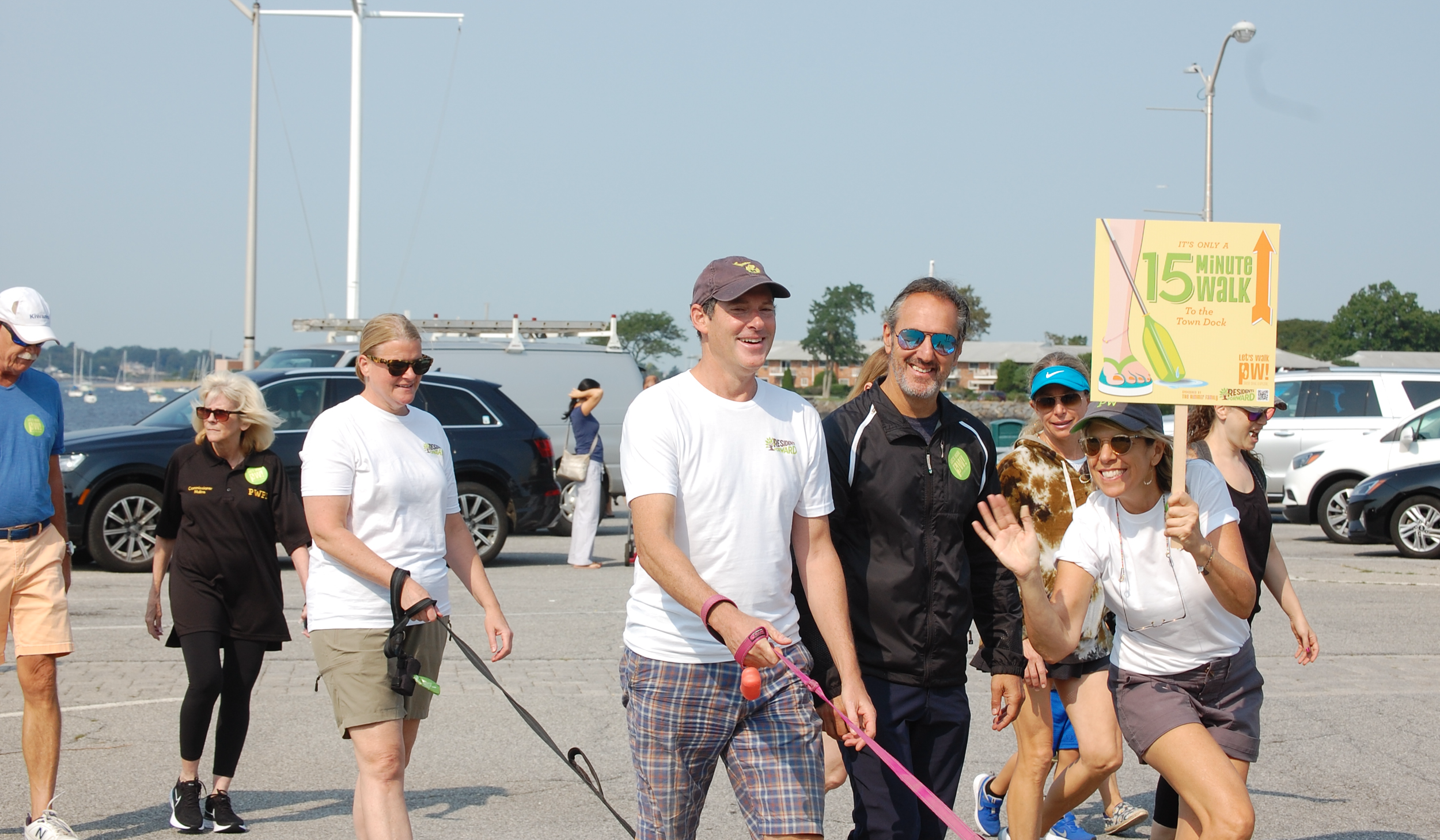 Group of people walking and smiling outdoors near water, some holding leashes and a woman holding a sign about a 15-minute walk to the town dock.