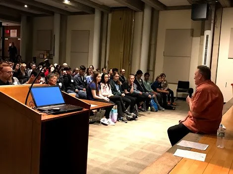 A man sitting on a stage speaking to an audience in a conference room, with a laptop, papers, and water bottles on a table.