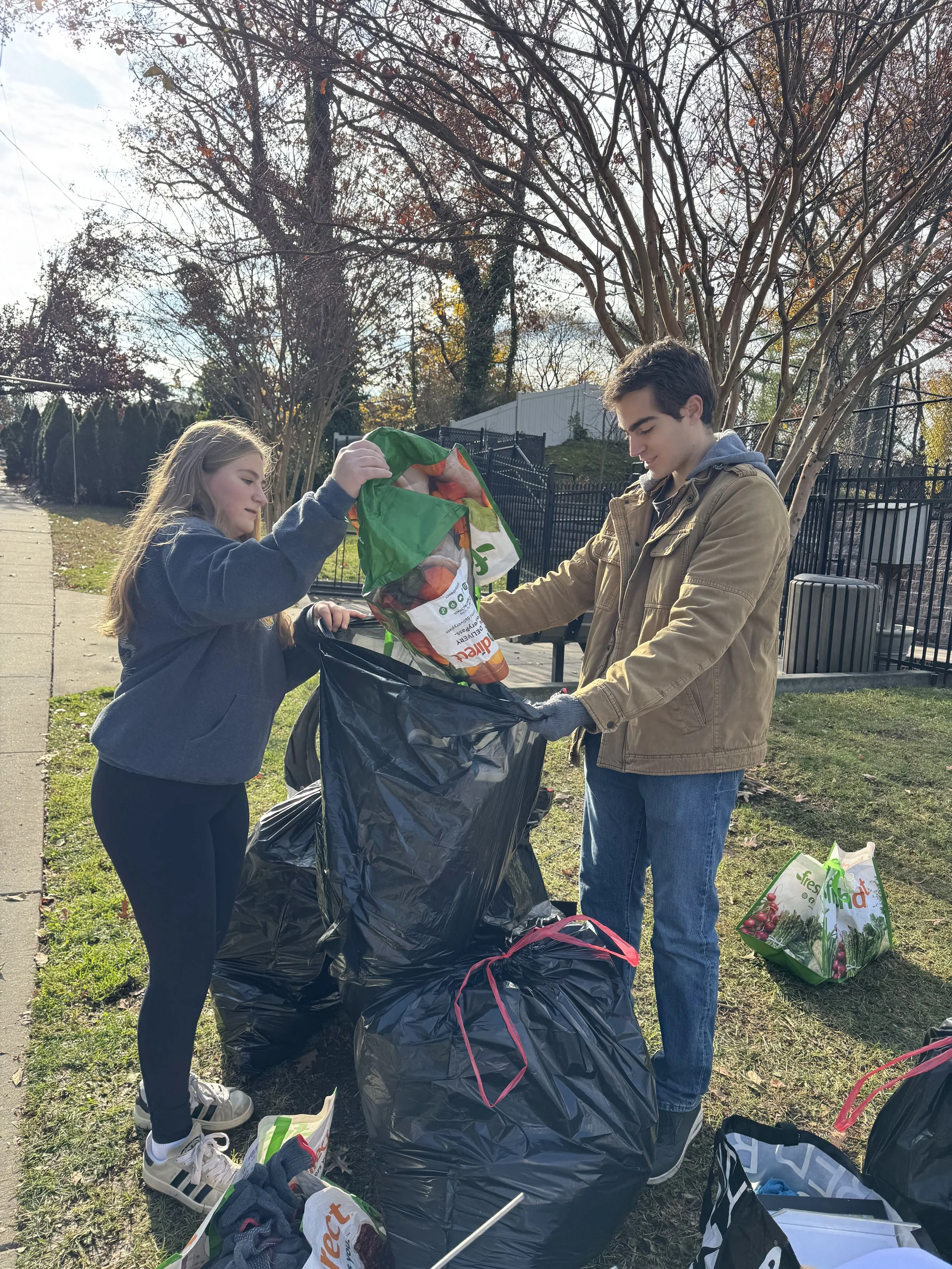 Two teenagers, a girl and a boy, collecting leaves or yard waste into large black trash bags outside in a park or neighborhood. The girl is holding a bag open, and the boy is placing leaves into it. Fall trees with bare branches and an iron fence are