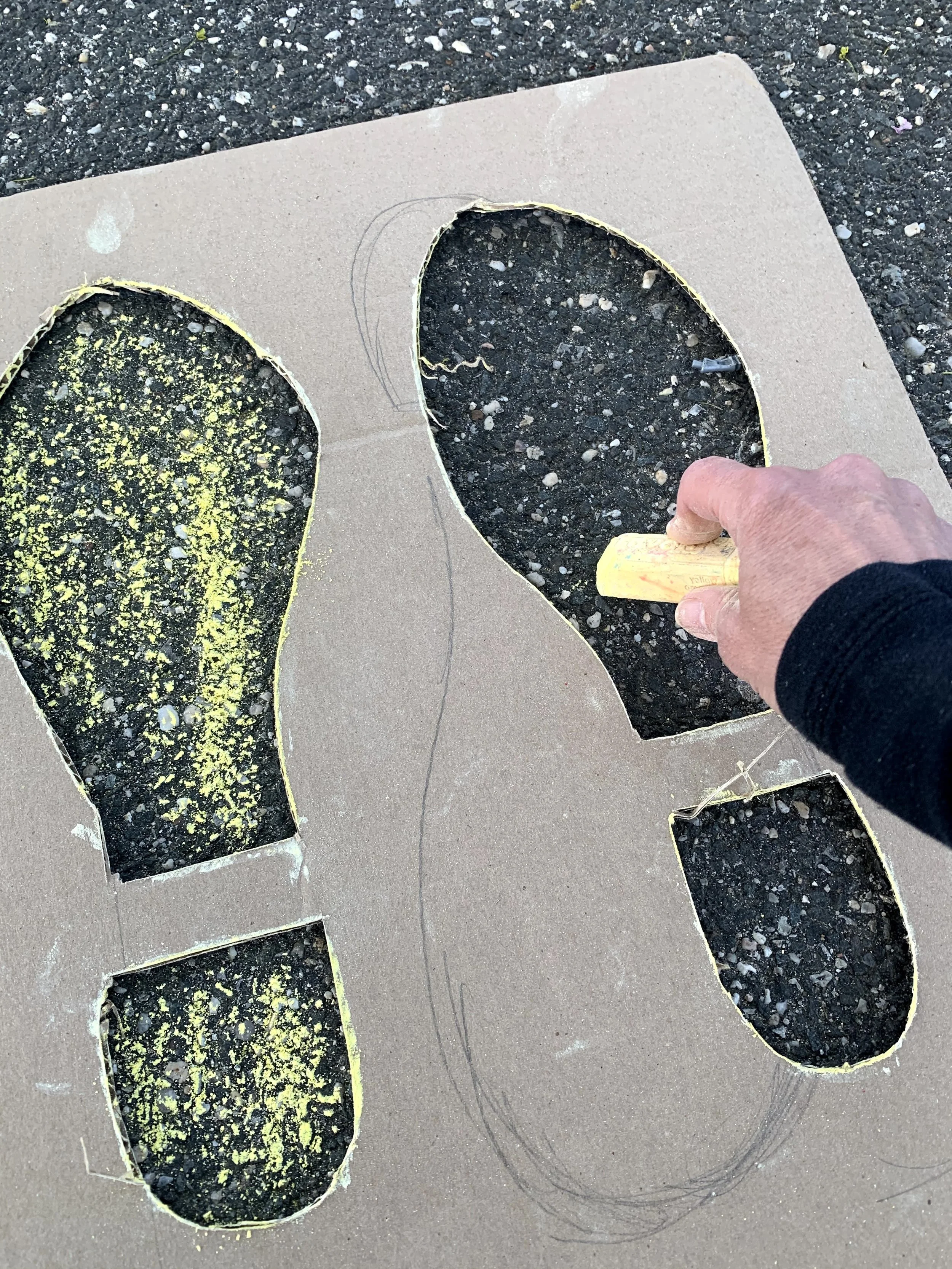 A person is tracing the outline of cut-out shoe shapes on a piece of cardboard with a yellow chalk in their right hand.