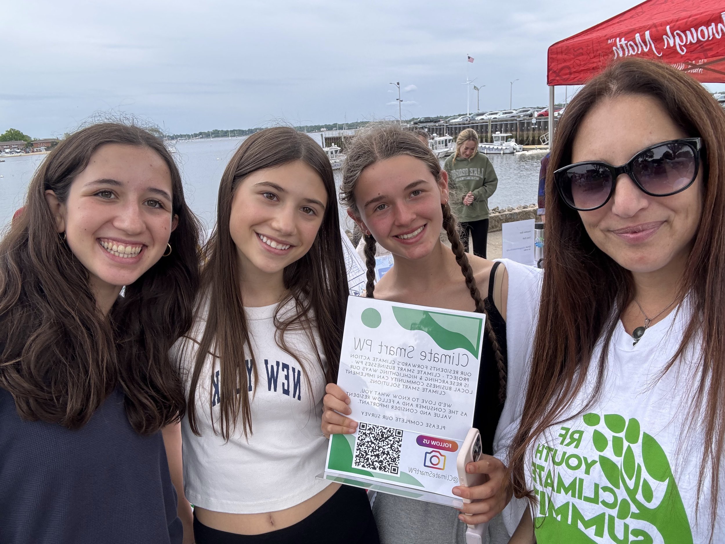 Four women smiling at the camera at an outdoor event near a body of water with boats and docks. One woman holds a flyer promoting environmental conservation.
