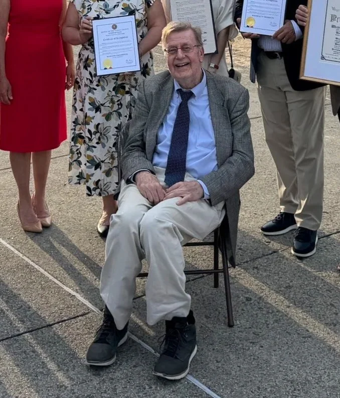 An elderly man wearing a gray blazer, light blue shirt, and black sneakers is sitting on a chair, smiling. Behind him, a group of people is standing, holding certificates, in an outdoor setting.
