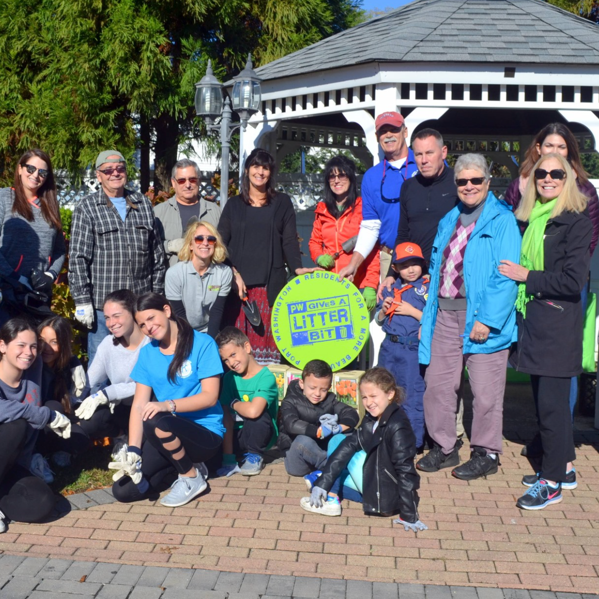 Group of people including children and adults participating in a community cleanup event, holding a sign that reads "PW GIVES A LITTER BIT", outdoors in a park with trees and a gazebo in the background on a sunny day.