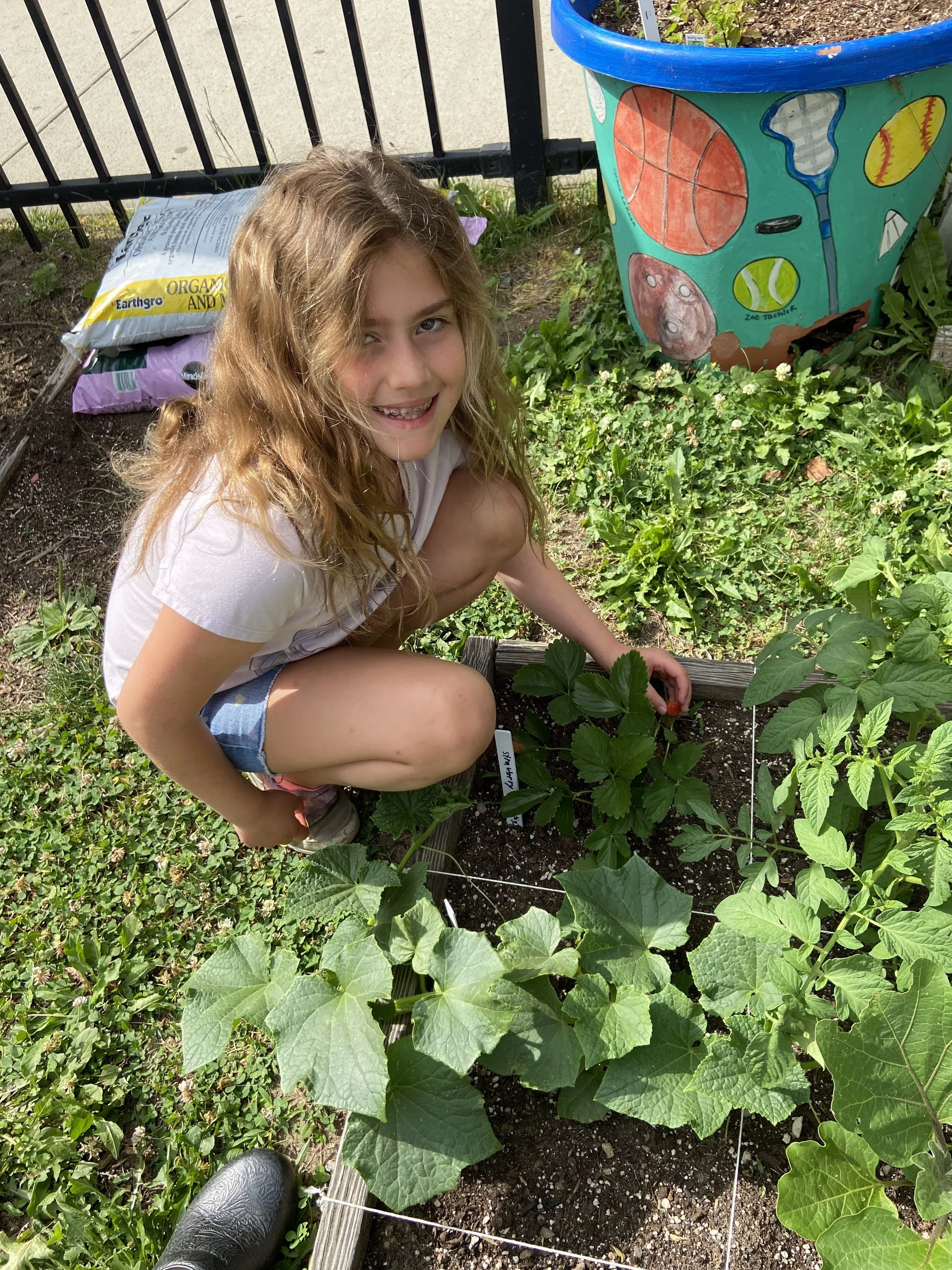 A young girl with curly hair and braces is crouching in a garden, smiling at the camera, next to a row of green plants. Behind her, there are bags of garden soil and a painted flower pot.