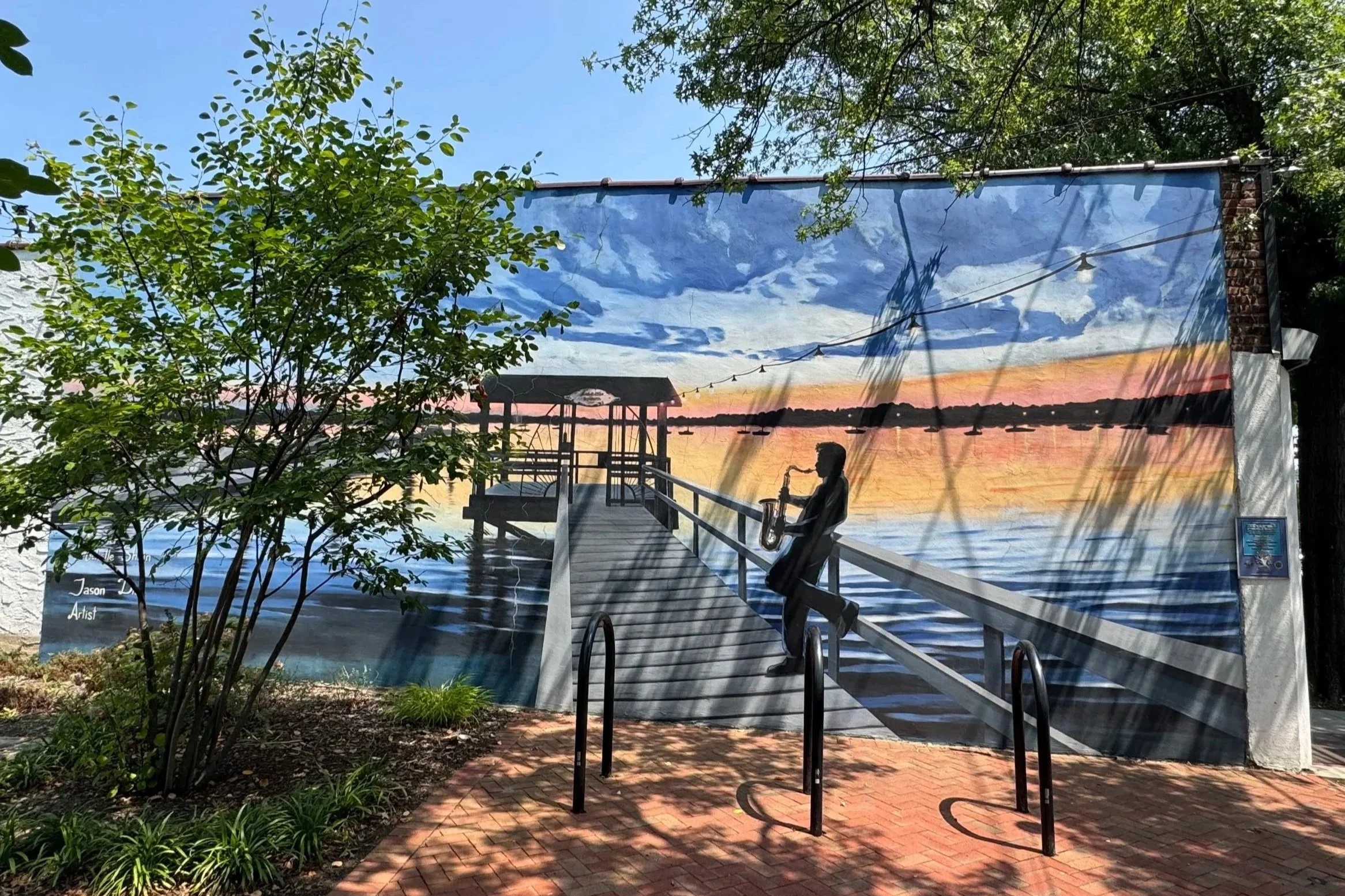 Titled 'Port Sunset' this wall mural depicts a wooden dock extending into a body of water at sunset, with a silhouette of a saxophone player on the dock, surrounded by trees and outdoor lighting.