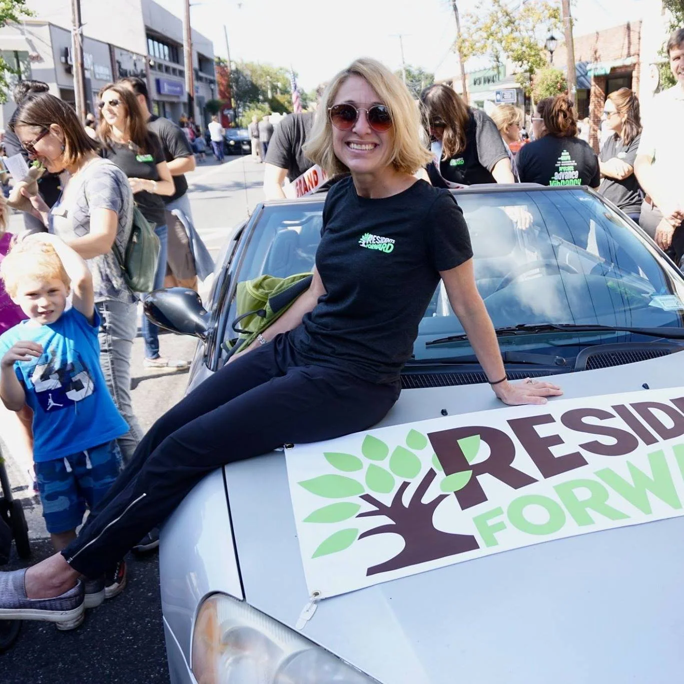 A woman with blonde hair wearing sunglasses and a black t-shirt that says 'President Bernie 2020' sitting on the hood of a silver car during a political rally or parade, with a crowd of people and storefronts in the background.