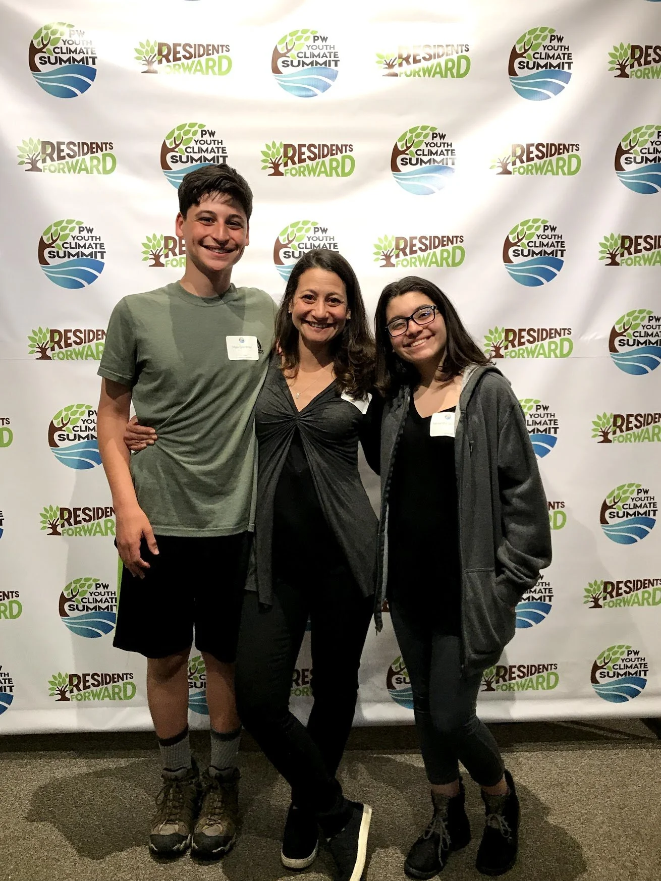 Three people standing together at an event, in front of a backdrop with logos including green leaves, a tree, water, and the text "RESIDENTS FORWARD" and "PW YOUTH CLIMATE SUMMIT." The person on the left is a young man with short dark hair, wearing a