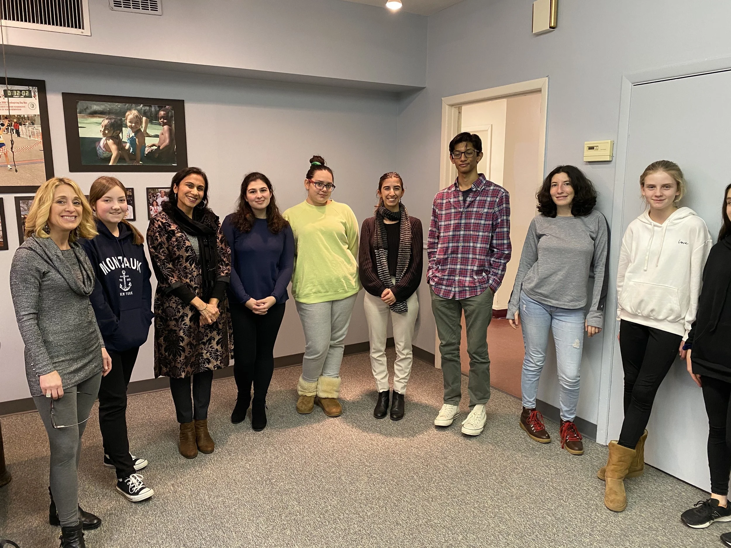 Group of ten people standing indoors, facing the camera, with a wall featuring framed photos in the background.