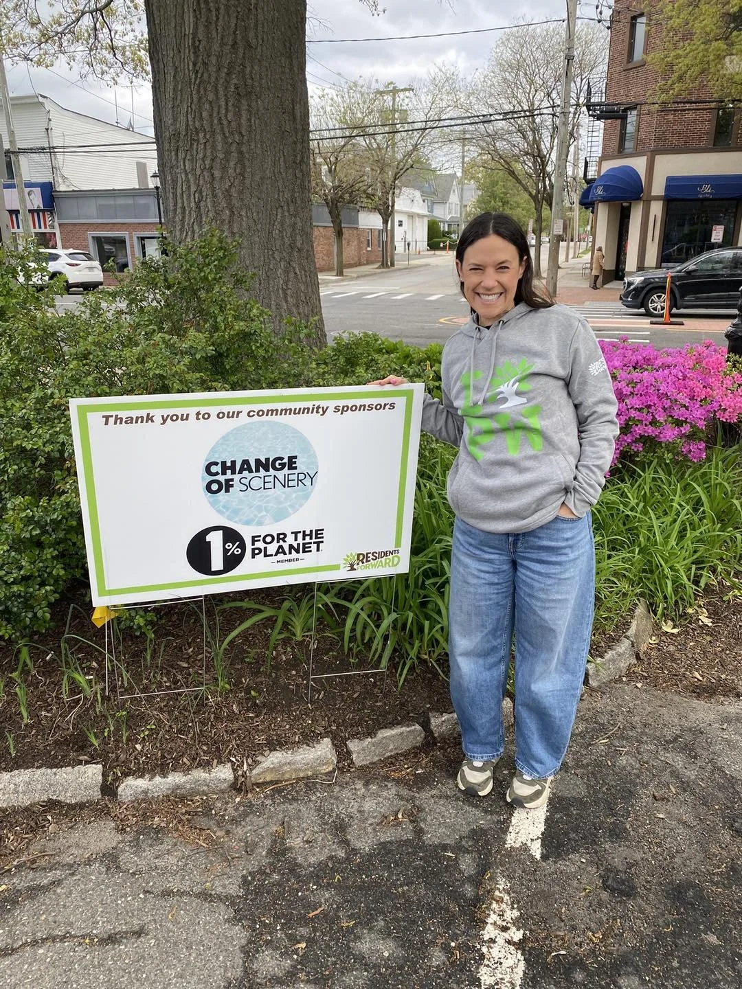 Woman standing outdoors, smiling, next to a sign that thanks community sponsors, with a background of trees, a building, and pink flowers.