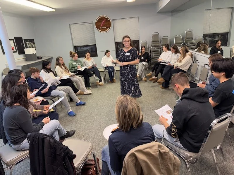 A woman standing in the center of a circle of seated young adults, speaking to a group in a room with windows, stacked chairs, and a clock on the wall.
