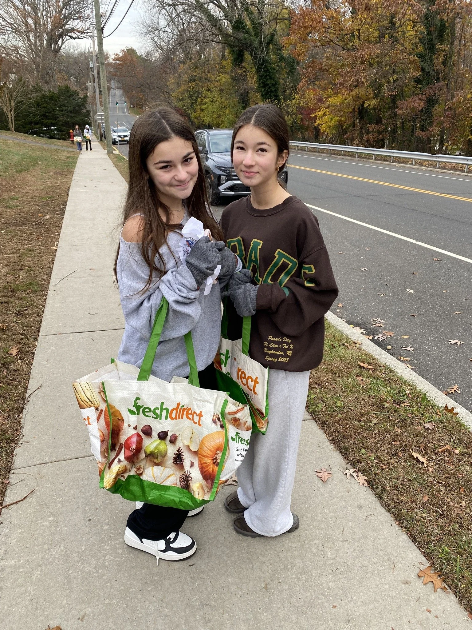 Two young girls standing on a sidewalk holding shopping bags, smiling, with a road and trees in the background.