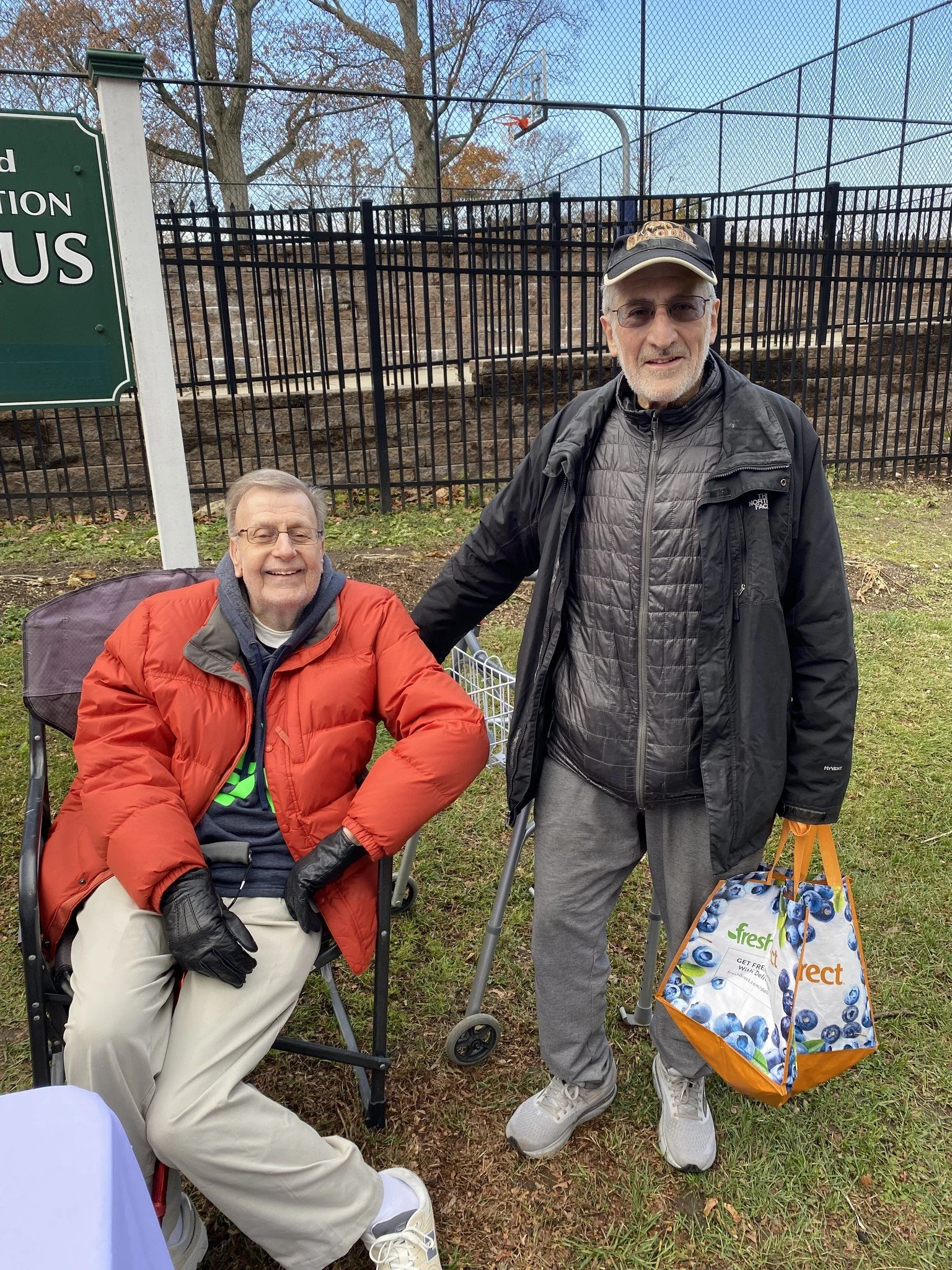Two older men outdoors near a fenced area, one sitting in a wheelchair wearing a red jacket and gloves, smiling, and the other standing with a walking aid holding a bag, both dressed warmly, with trees and a basketball hoop in the background.