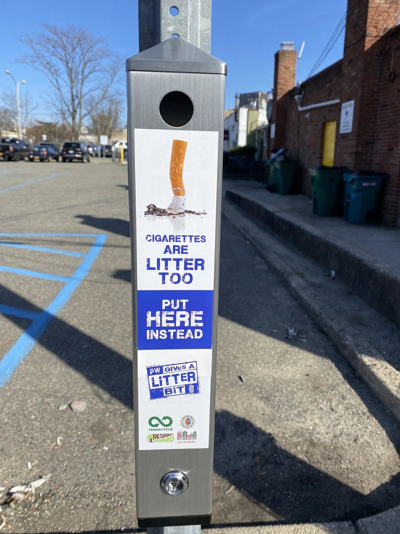 A public trash bin with a sign that encourages cigarette smokers to dispose of their cigarette butts properly. The sign features a picture of a cigarette butt on the ground and text that reads, "Cigarettes are litter too. Put here instead." Additional social and environmental messages promote recycling and community responsibility.