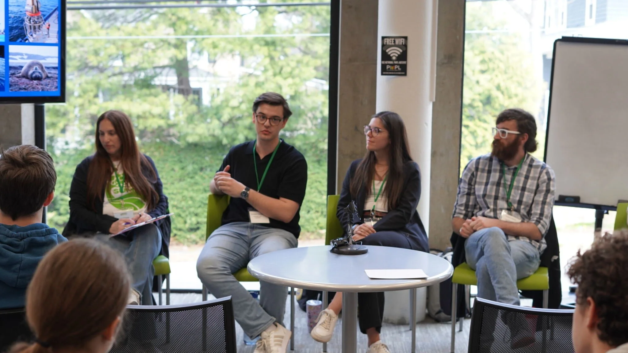 Four speakers seated on stage in a panel discussion at a conference, with a large window and trees in the background, and an audience in front.