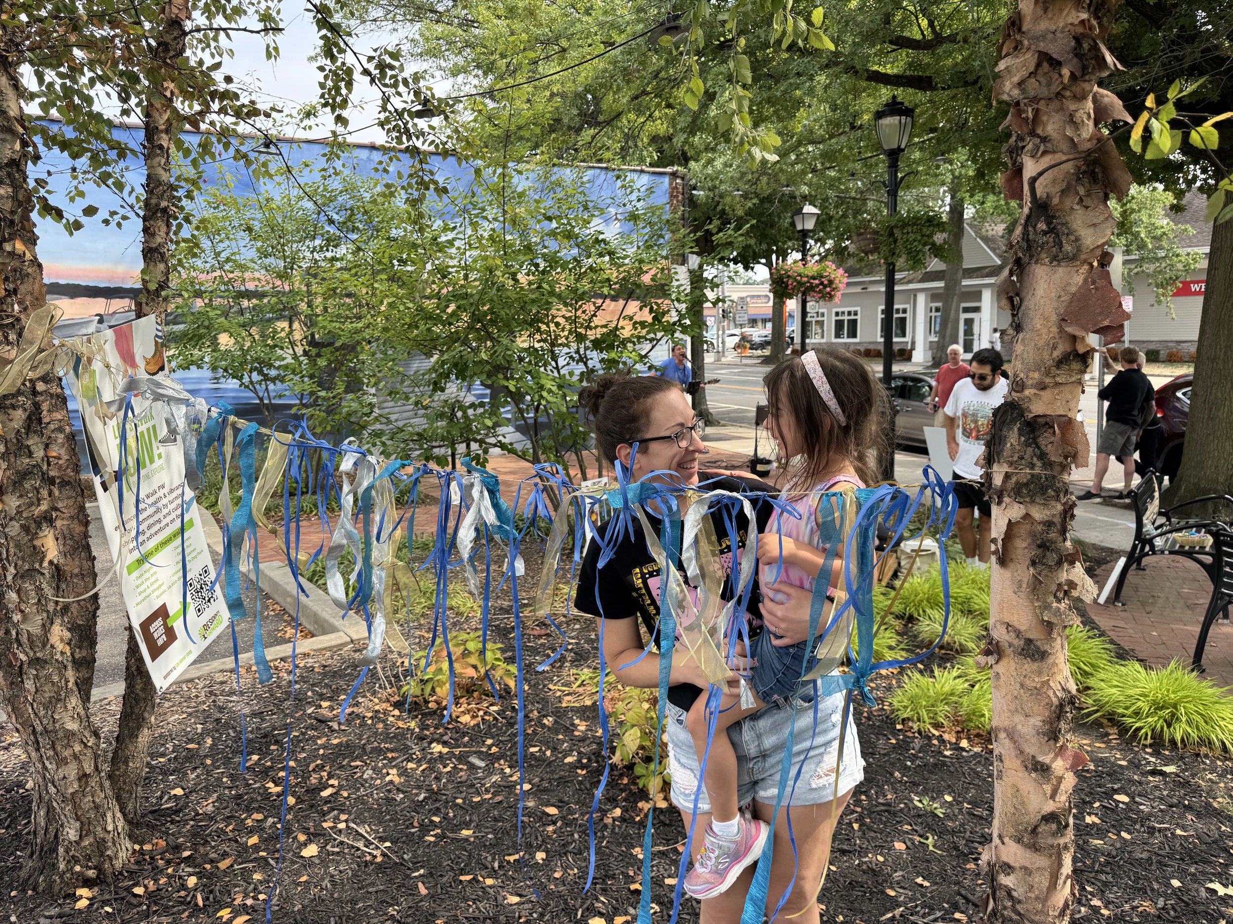 A woman holding a young girl and smiling at each other in front of a decorated tree with blue and silver ribbons, in a park setting on a sidewalk with people walking and sitting nearby.
