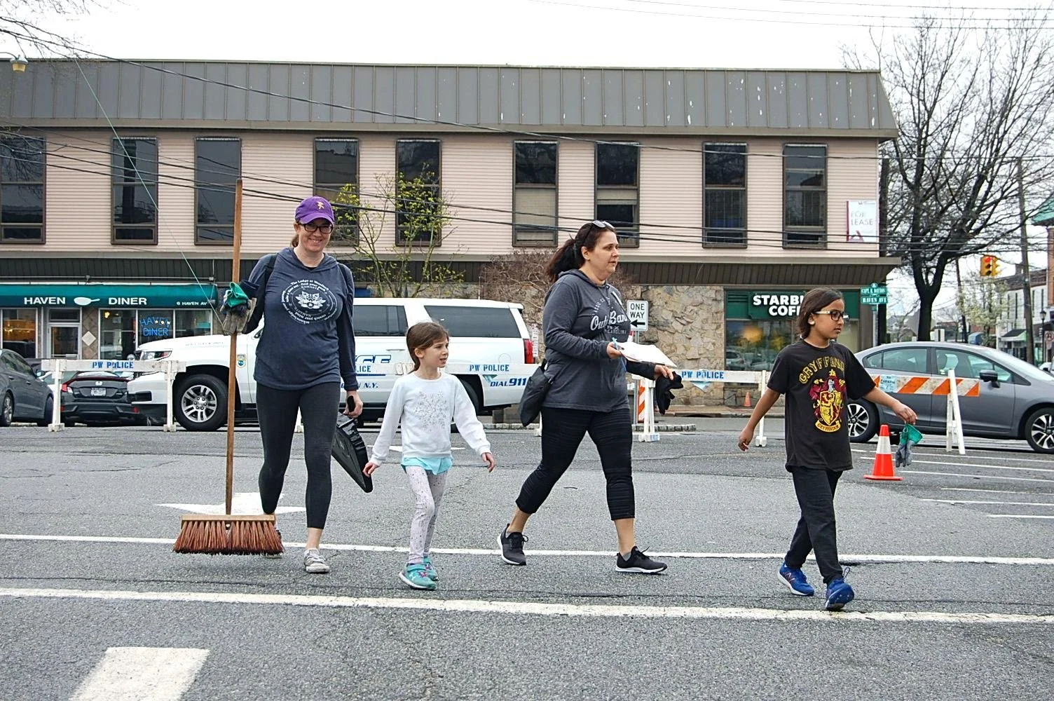 Five women and two children walking across a street in an urban area, with parked cars, a police vehicle, and storefronts in the background, some carrying cleaning tools.