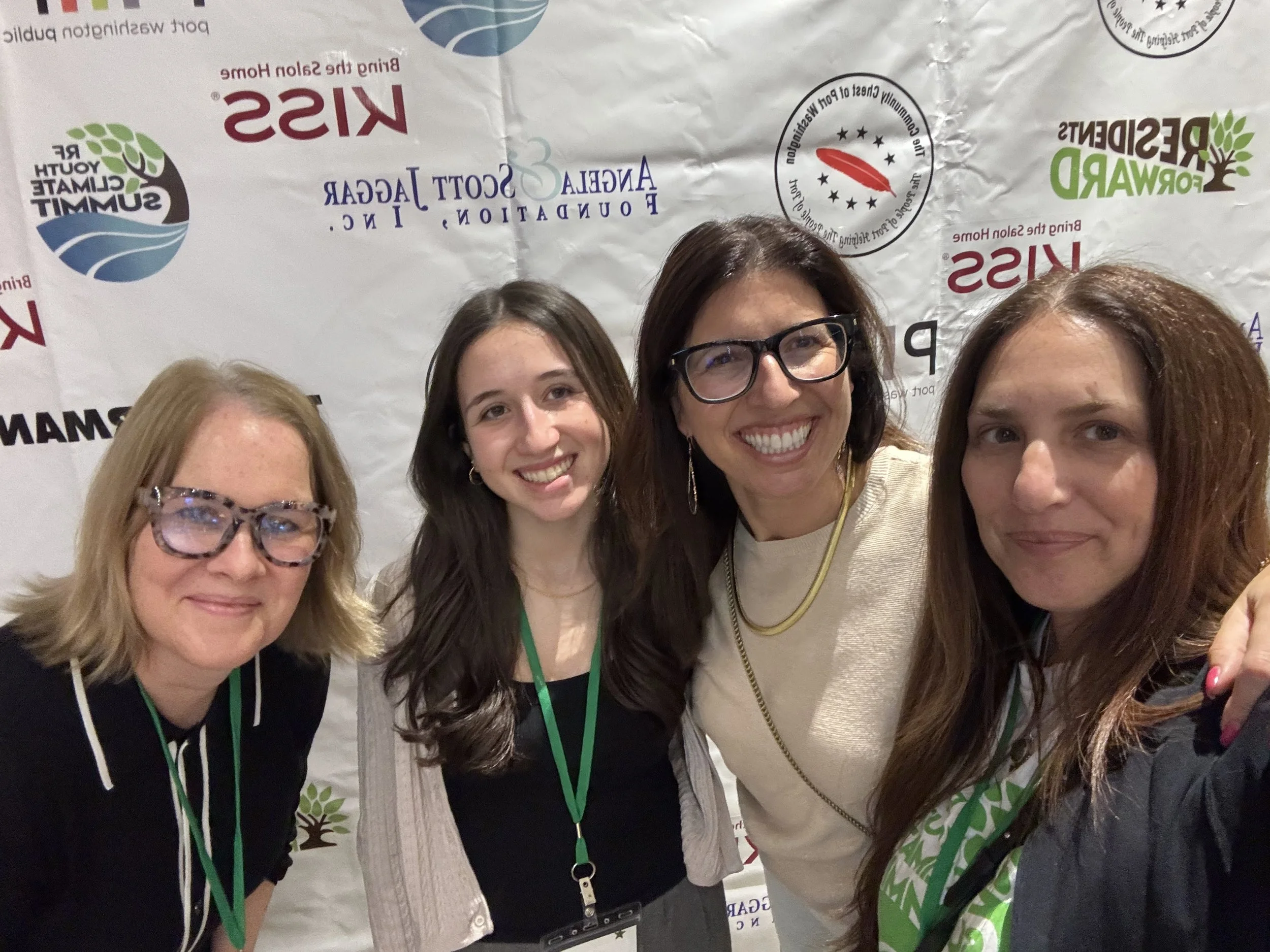 Four women taking a group selfie in front of a backdrop with logos and text related to climate and environmental initiatives. They are smiling and wearing conference badges.