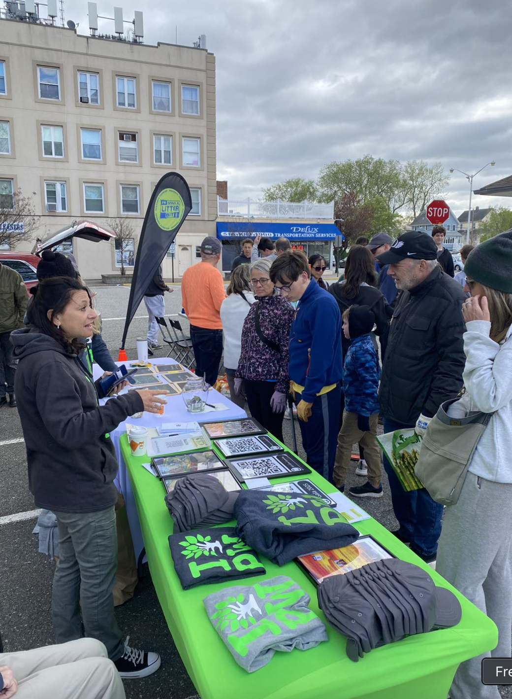 People standing in line at a recycling and environmental awareness event outdoors, with a table displaying informational materials, merchandise with eco-friendly messages, and QR codes, in an urban setting under cloudy skies.