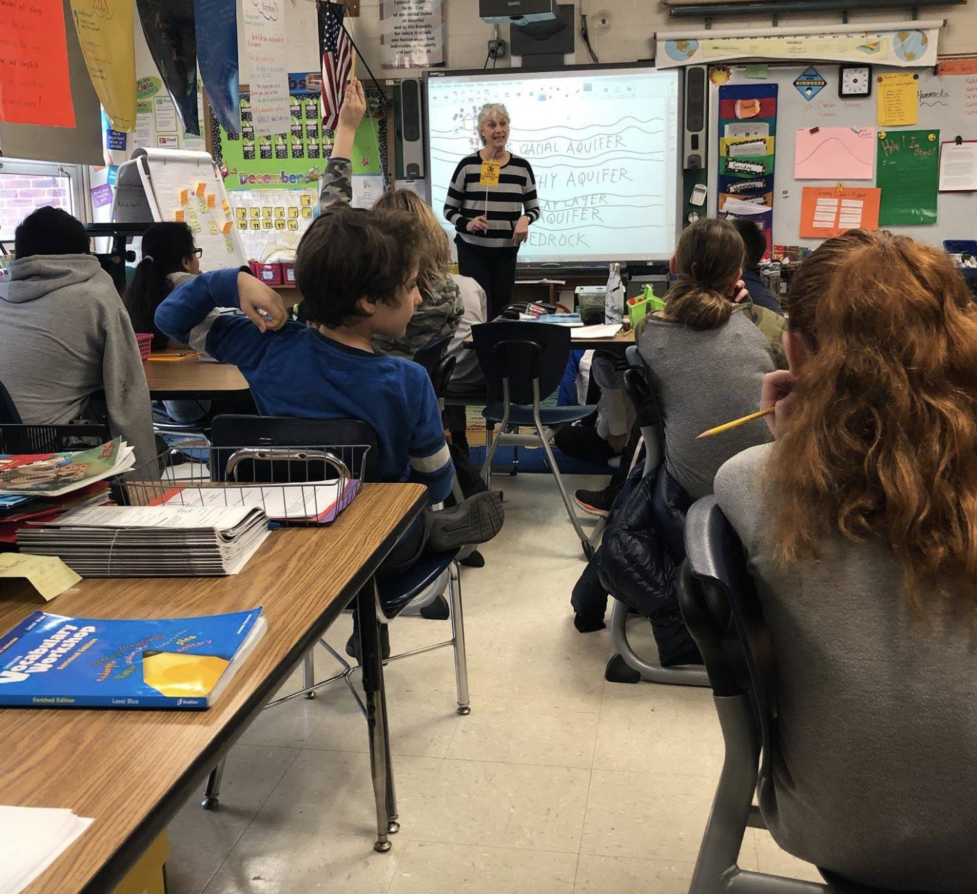 A classroom filled with students sitting at desks, facing a teacher at the front who is writing on a whiteboard. Students have books and supplies on their desks, some are taking notes, and one student has their hand raised.
