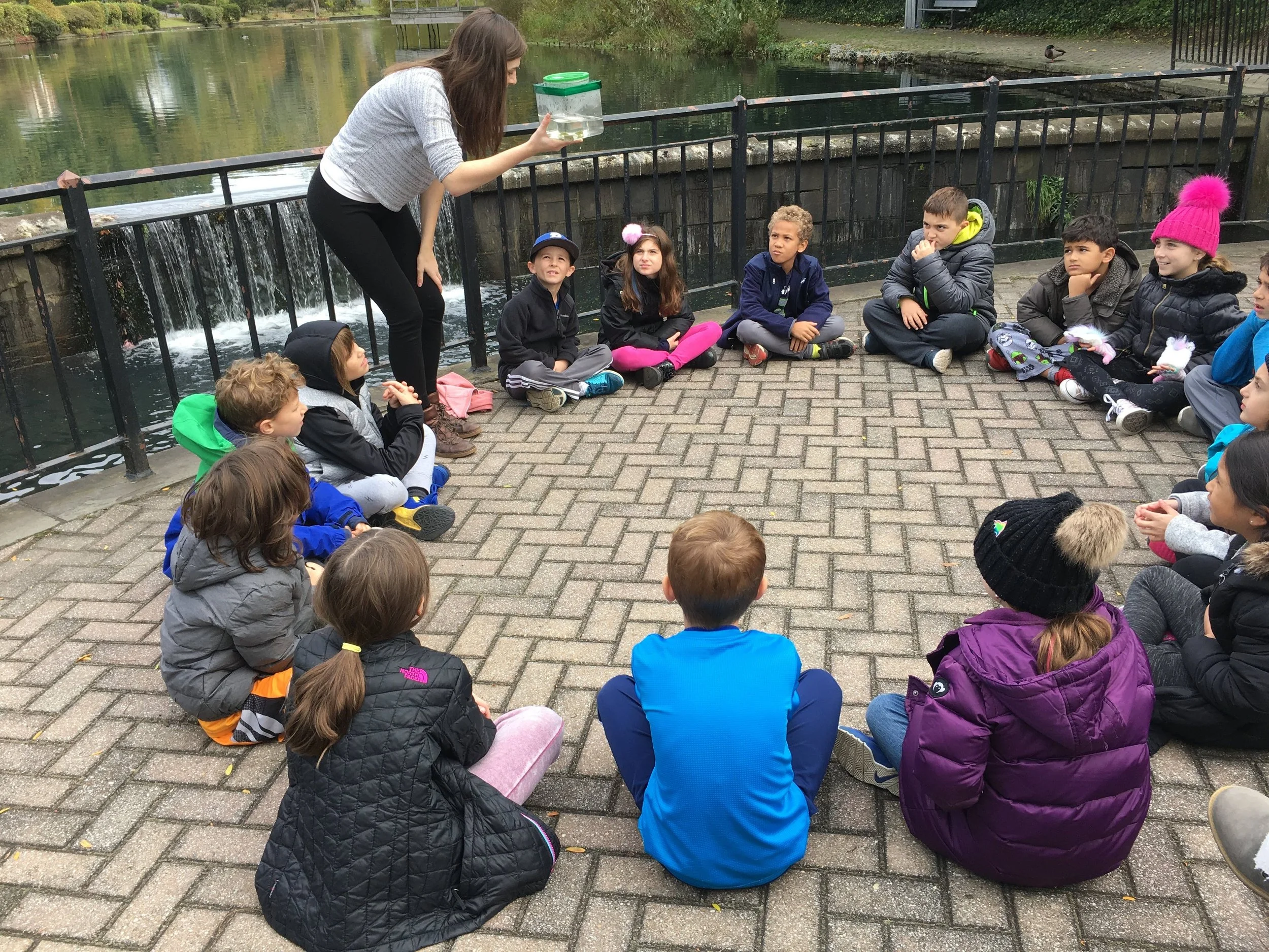 A group of children sitting in a circle on a paved area by a river, listening to a woman showing them insects or small creatures in a container.