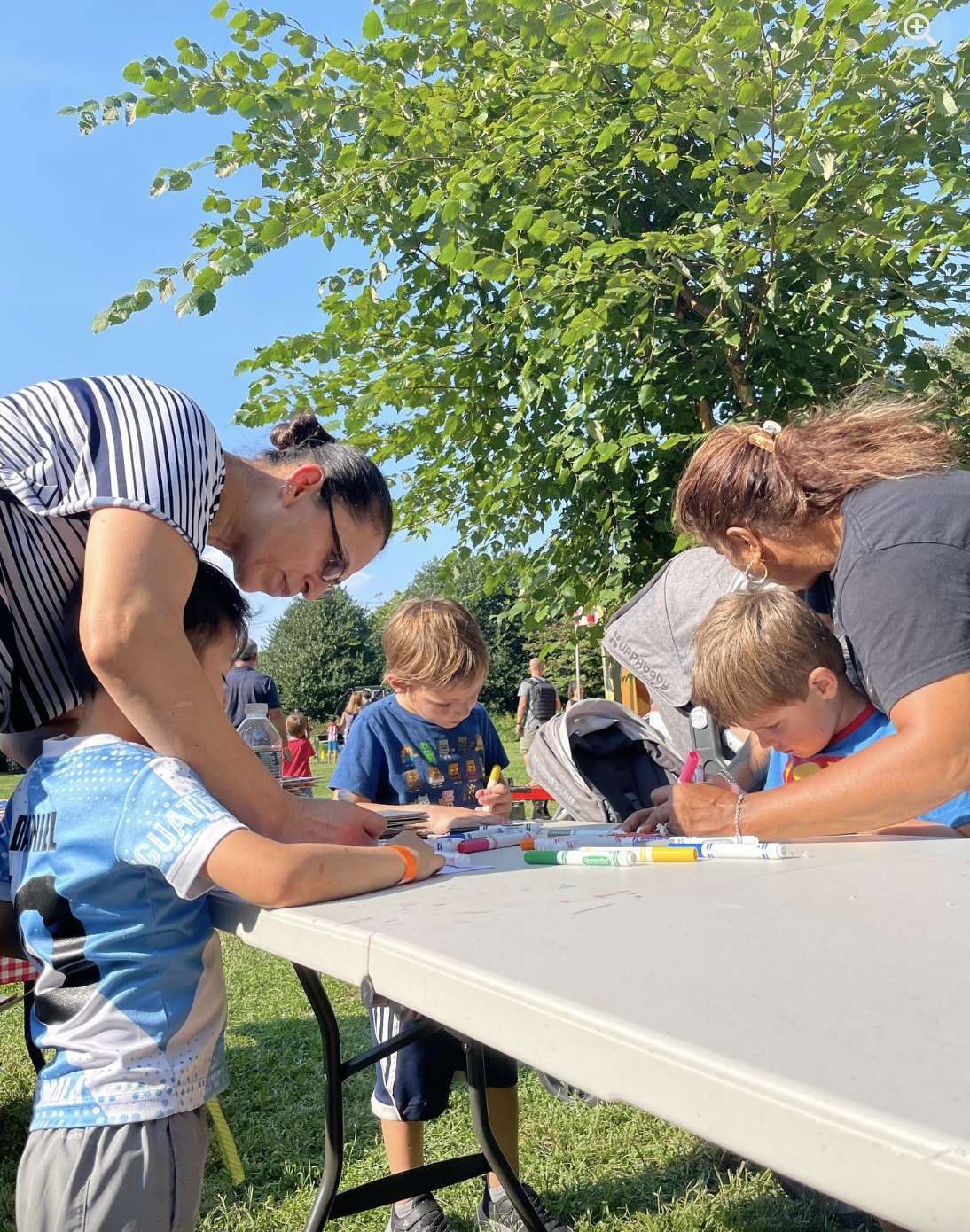 Children and adults gather around a table outdoors on a sunny day, drawing and coloring with markers, with green trees and a blue sky in the background.