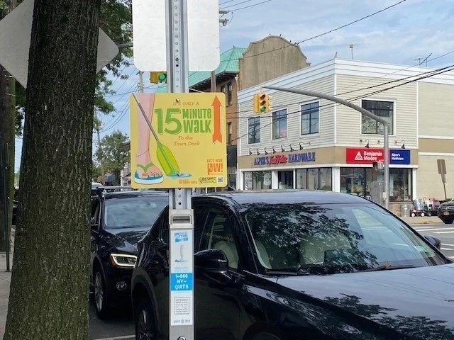 Street scene with cars parked along the curb, a sign indicating a 15-minute walk to the town dock, and buildings including a pharmacy, a barbershop, and a shop with a red awning in a small downtown area.
