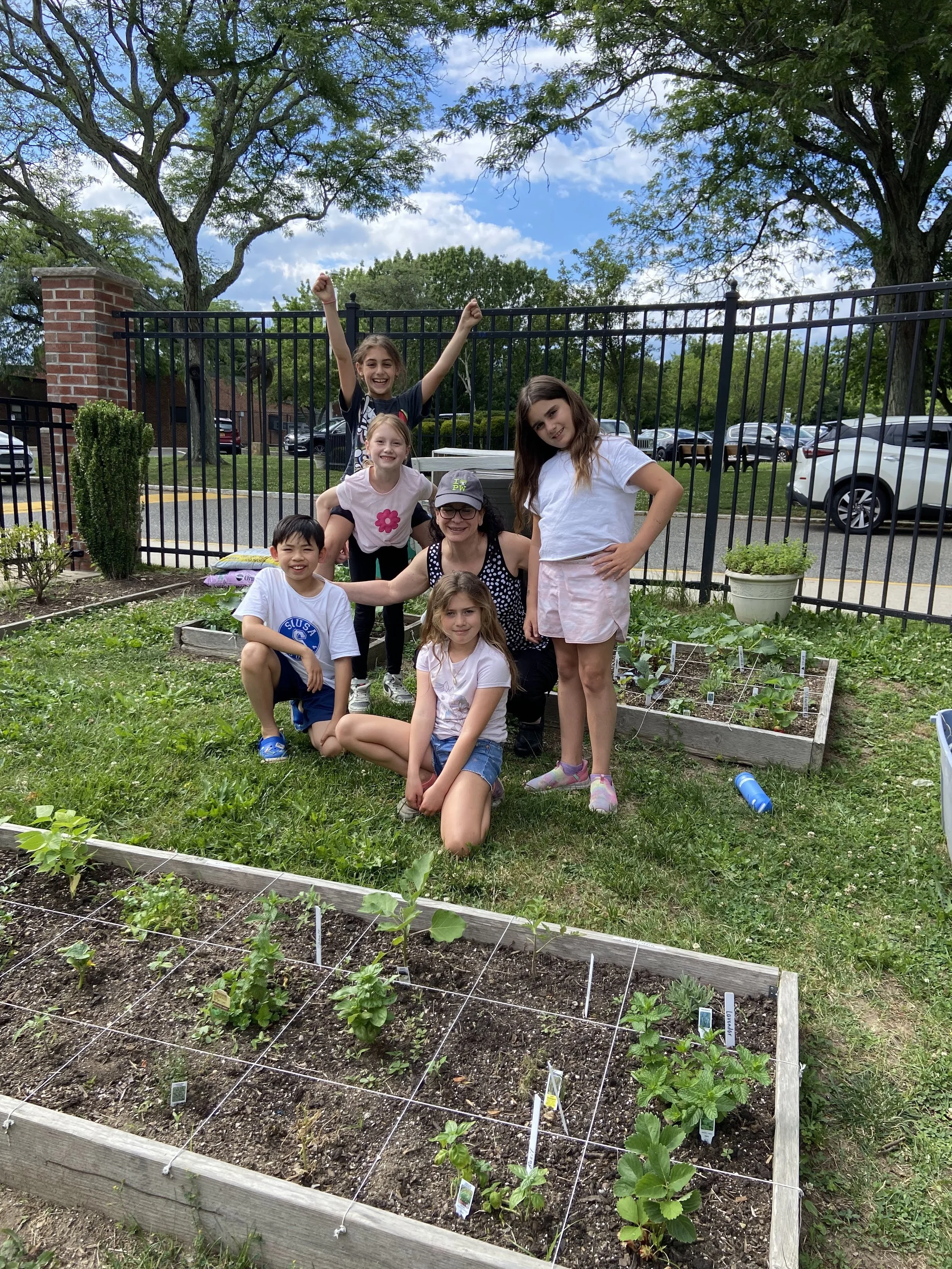 Group of children and a woman posing in a garden with vegetable beds and a fence behind them, under a partly cloudy sky.