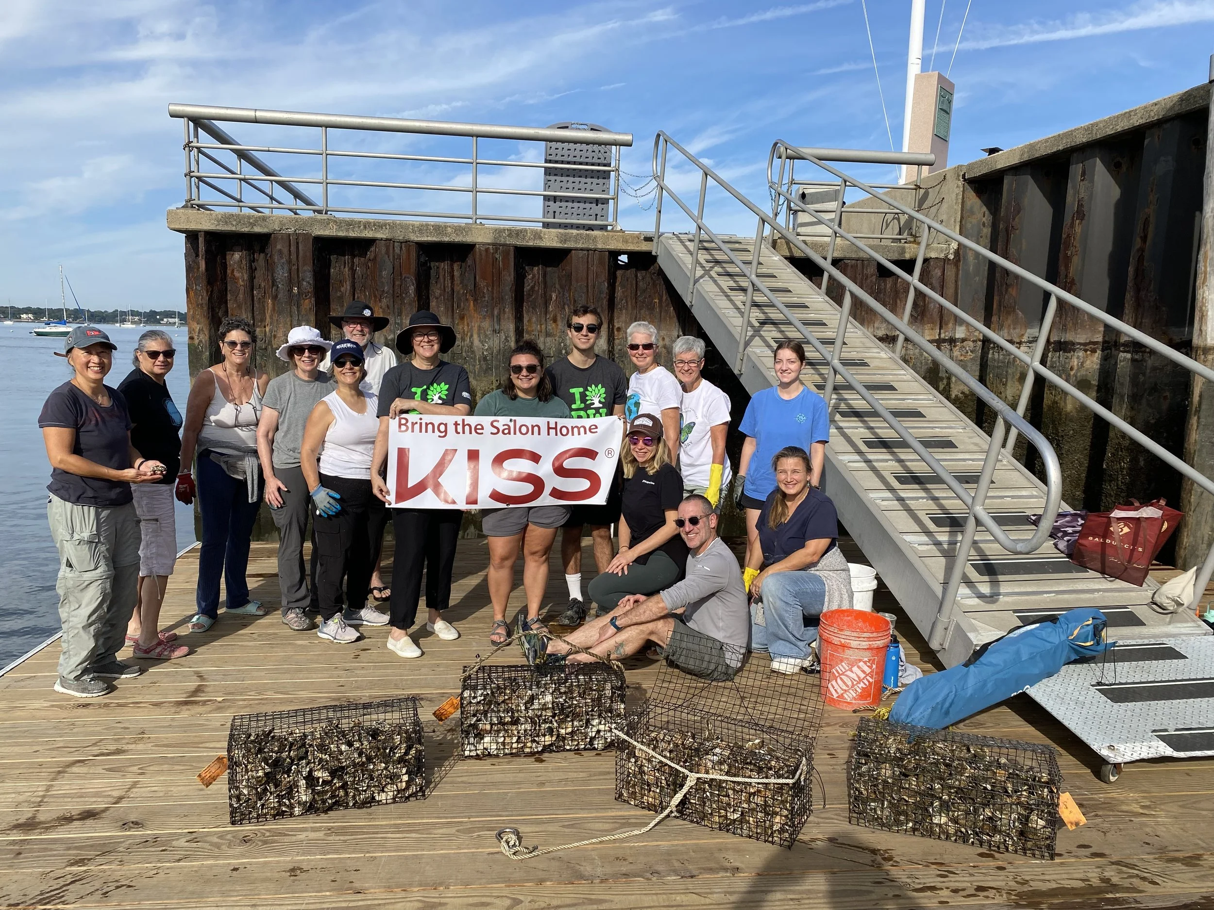 Group of people holding a banner that says "Bring the Salon Home KISS" on a dock near the water, with lobster traps and supplies in the foreground and boats on the water in the background.